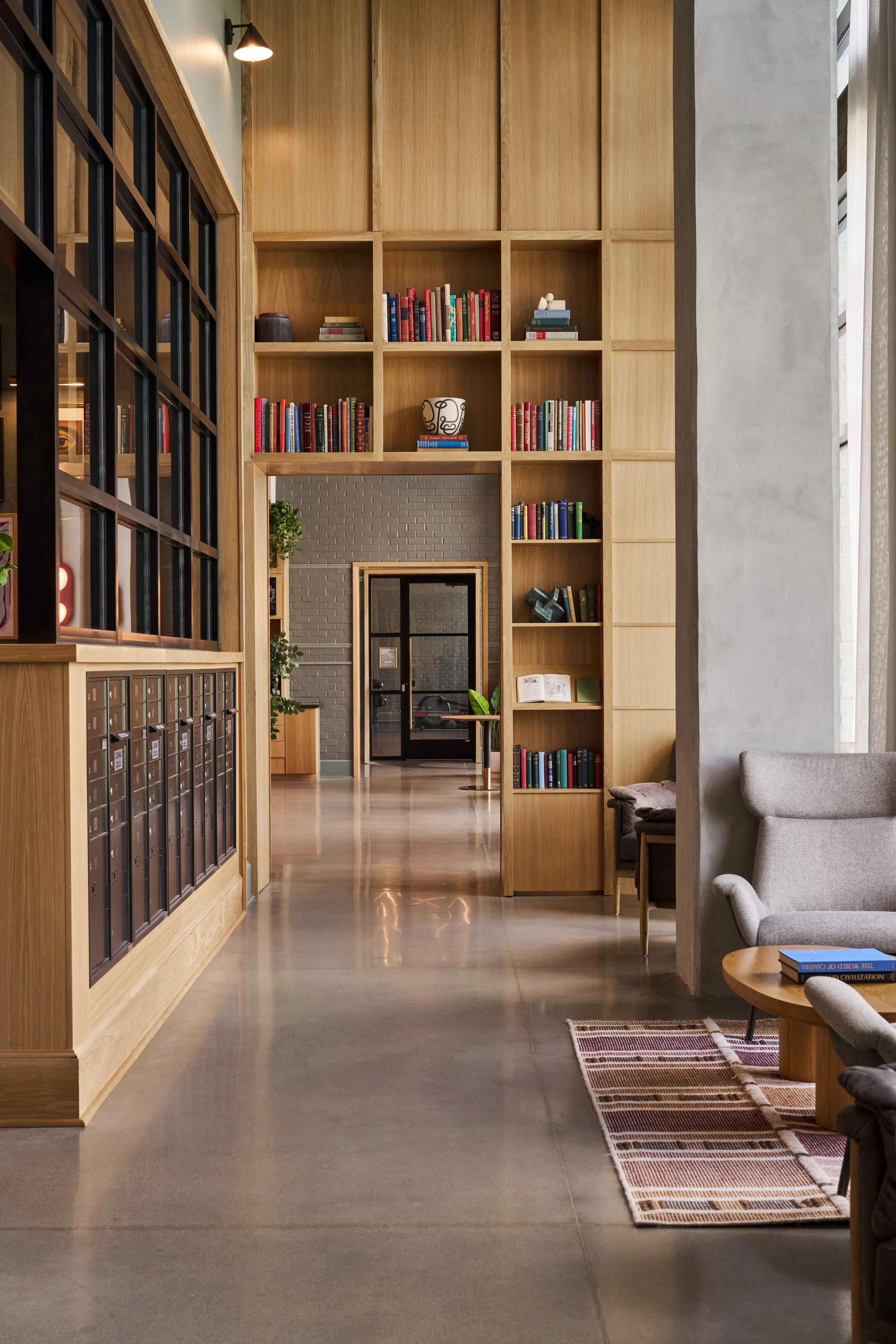 Interior of a modern library or lounge area with wooden bookshelves, a grey armchair, a small side table with books, a patterned rug, and large windows providing natural light.