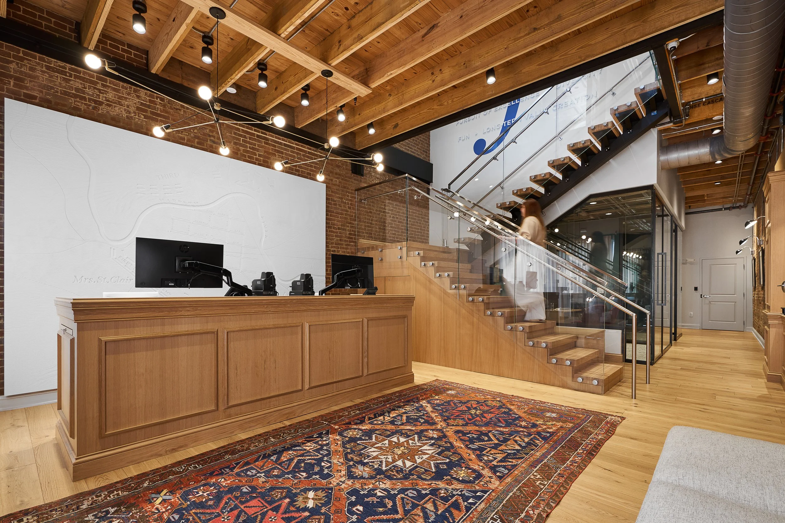 Interior of a modern office with wooden accents, exposed brick wall, a reception desk, computer, staircase with glass railing, and a decorative rug.