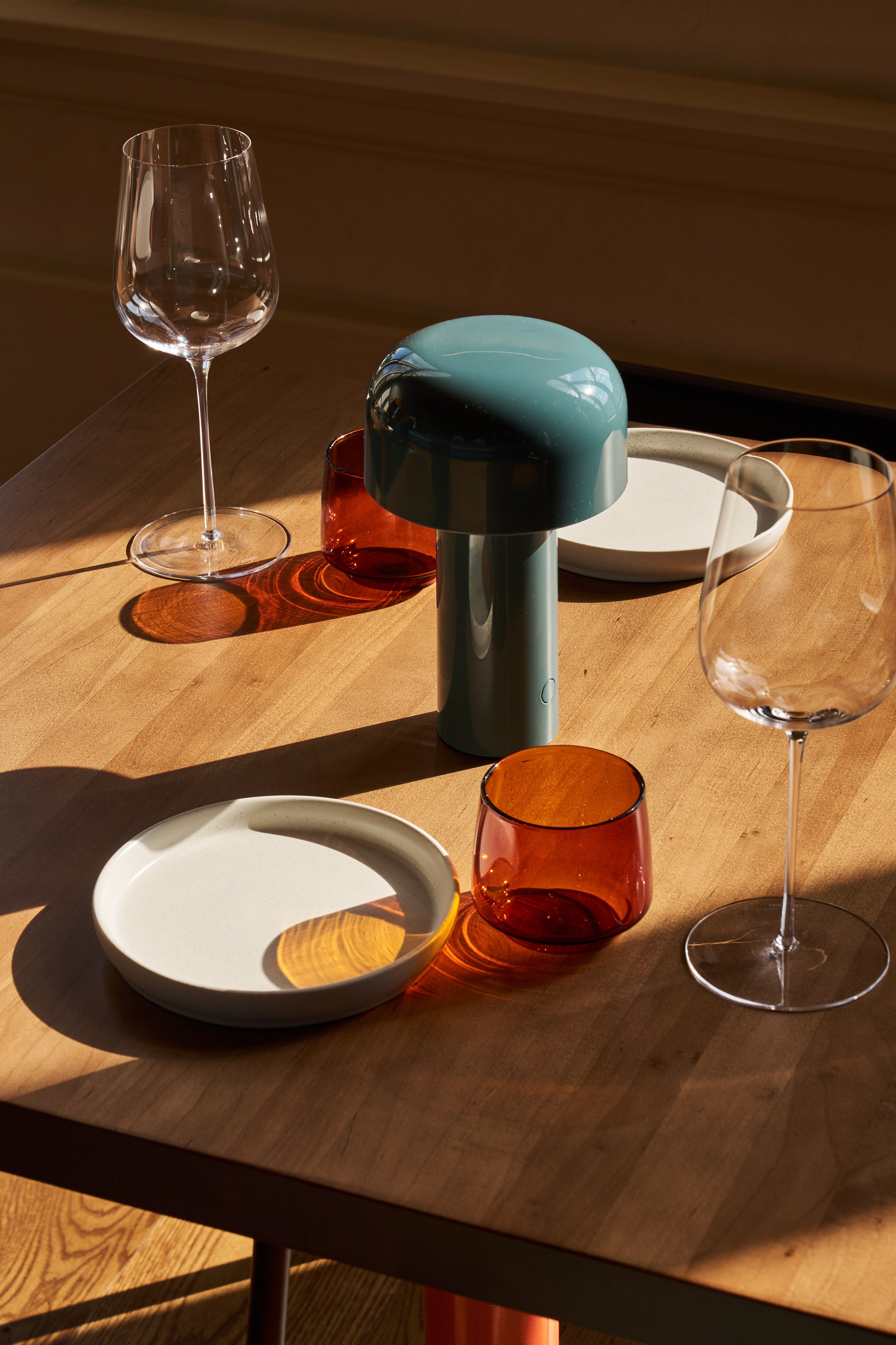 A wooden dining table with a transparent wine glass, a small amber glass, a white ceramic dish, a red glass, and a blue lamp with a grey base. Sunlight casts shadows on the table.