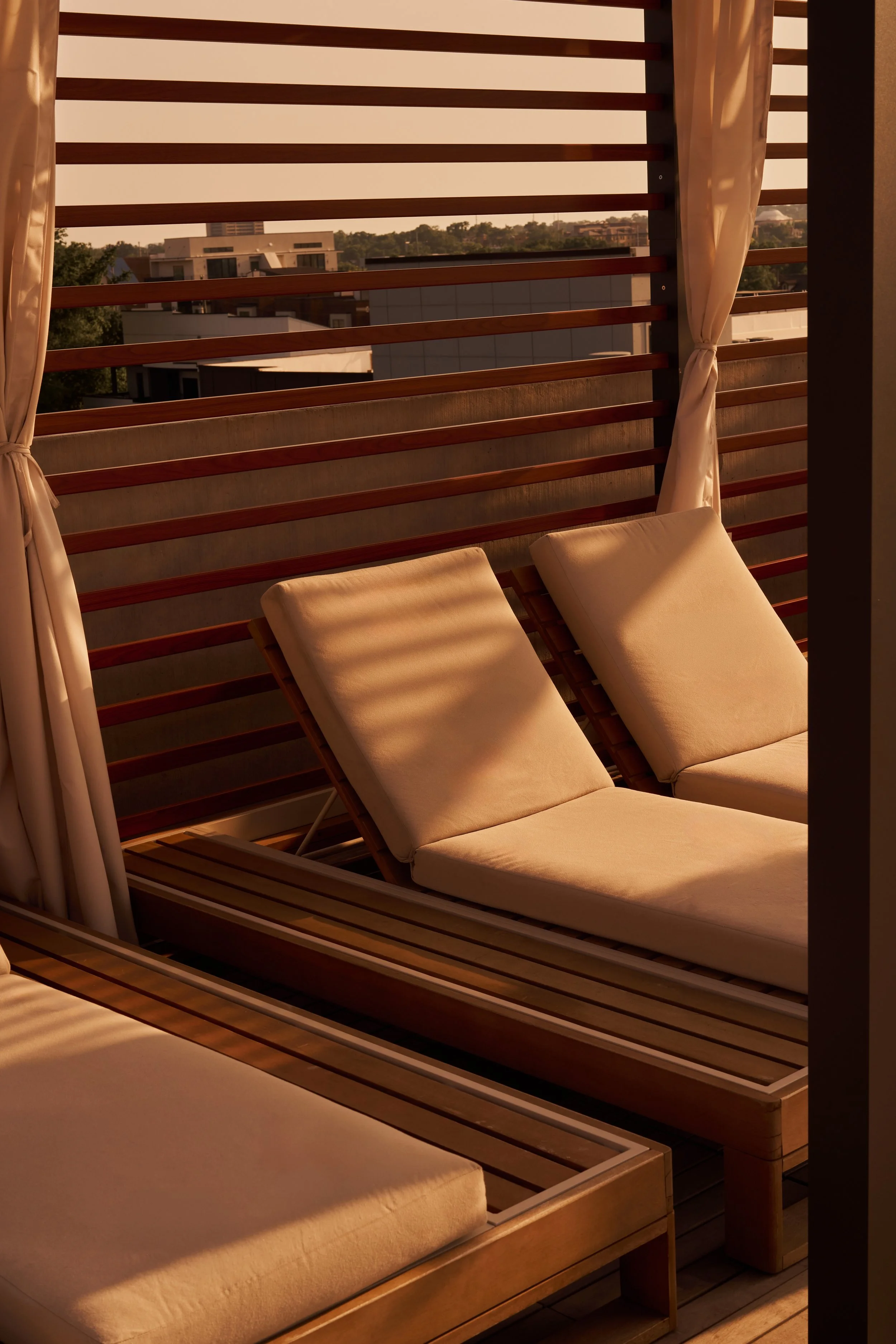 Two lounge chairs with cream cushions on a wooden deck, overlooking a cityscape with modern buildings and greenery in the background, during sunset.