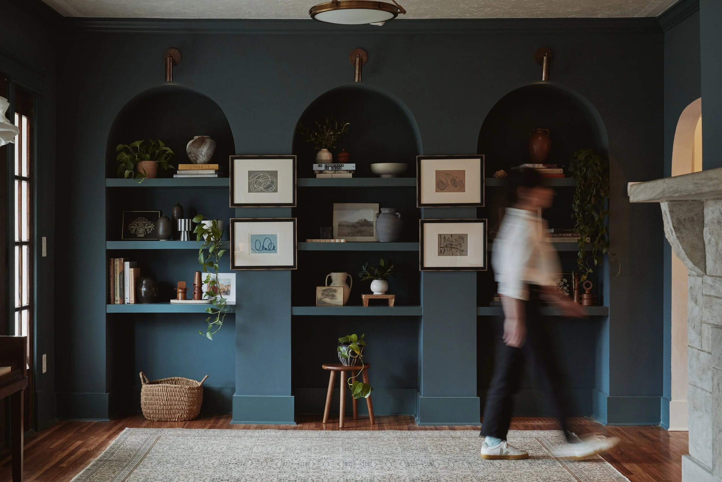 A person walking past a blue built-in bookshelf with decorative objects, framed artwork, and books in a living room with wooden flooring and a patterned rug.