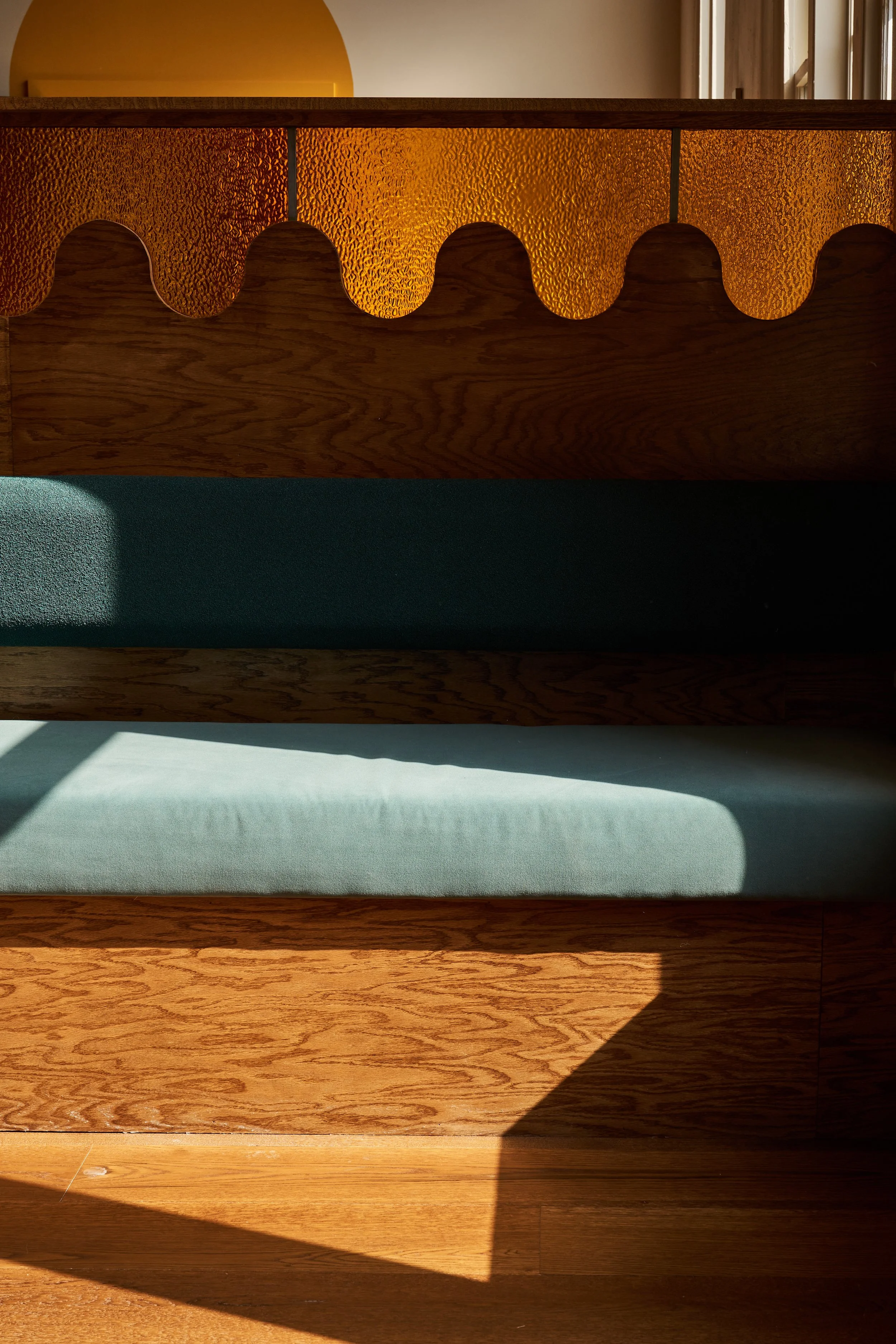 Close-up of a wooden cabinet with a textured, orange backlit glass panel featuring scalloped edges, placed against a wall with a window and sunlight casting shadows on the floor.