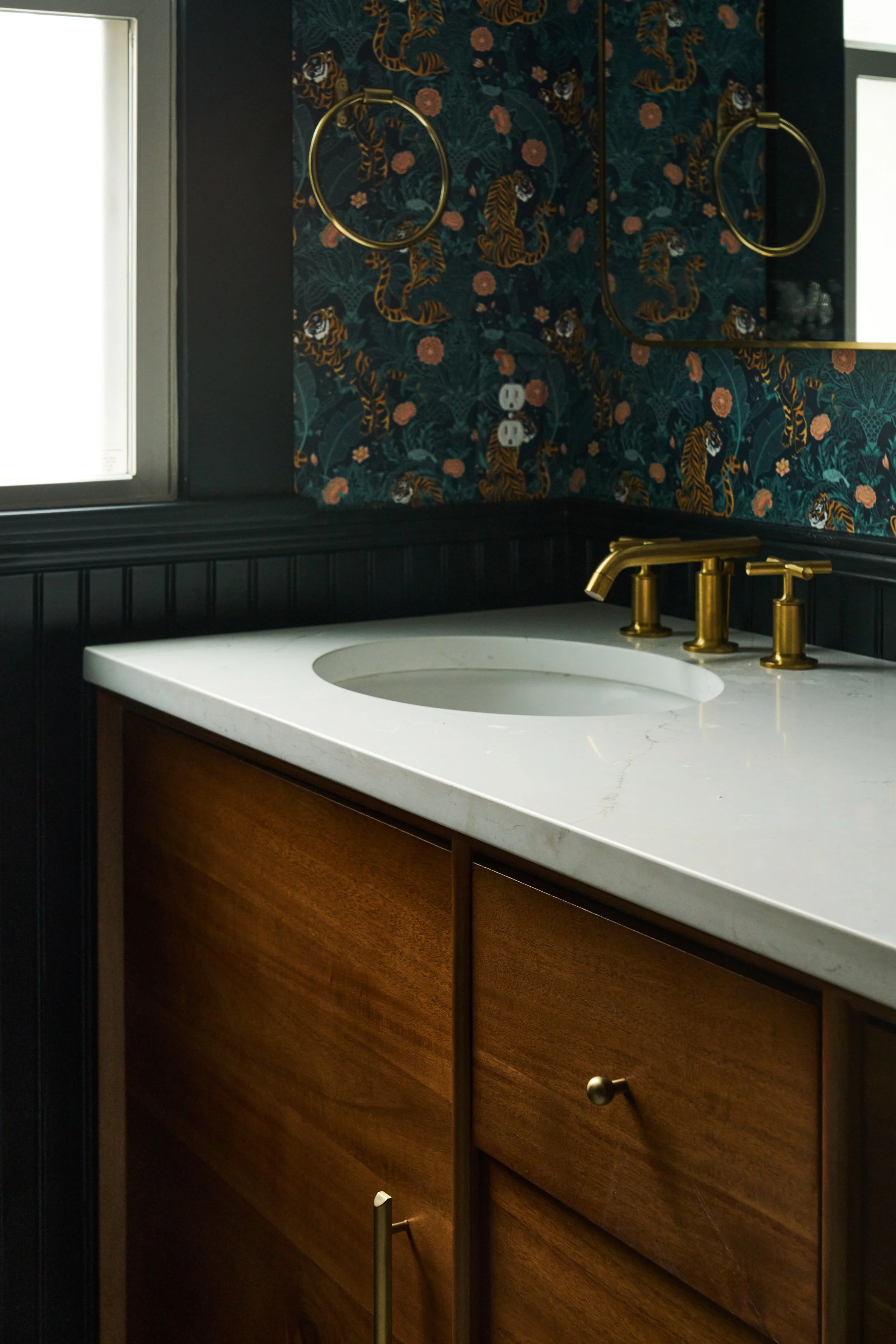 Bathroom vanity with a white countertop, a built-in sink, gold faucet, dark wooden cabinet, patterned wallpaper with tigers and flowers, large mirror, window, and double electrical outlets.