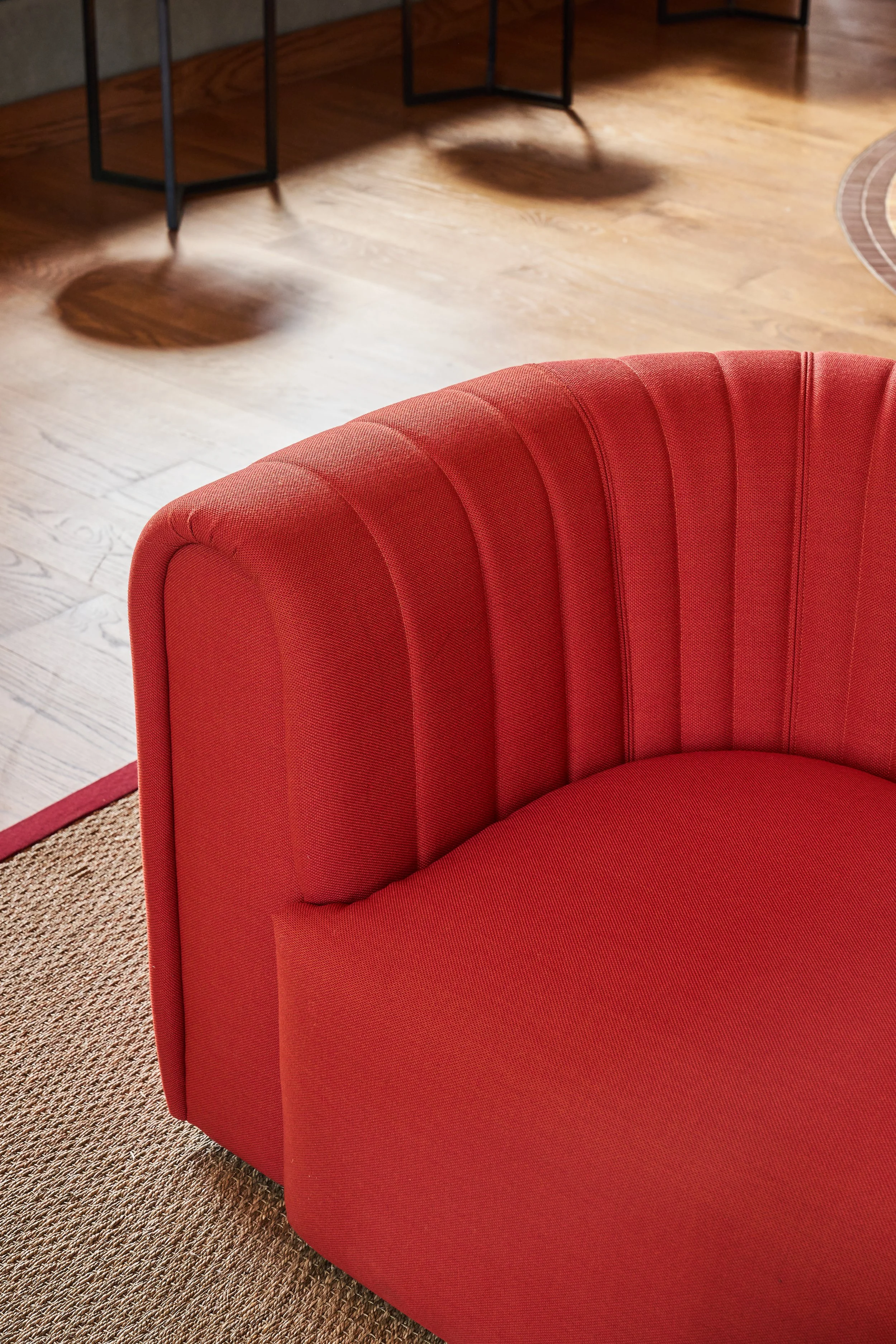 Close-up of a red upholstered chair with vertical stitching, on a beige textured carpet in a room with wooden flooring and black metal furniture.