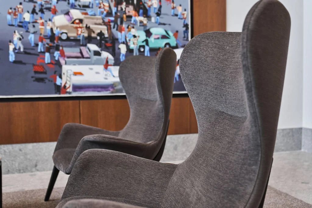 Two gray armchairs with high backs in a waiting area, with a colorful urban scene painting on the wall behind them.