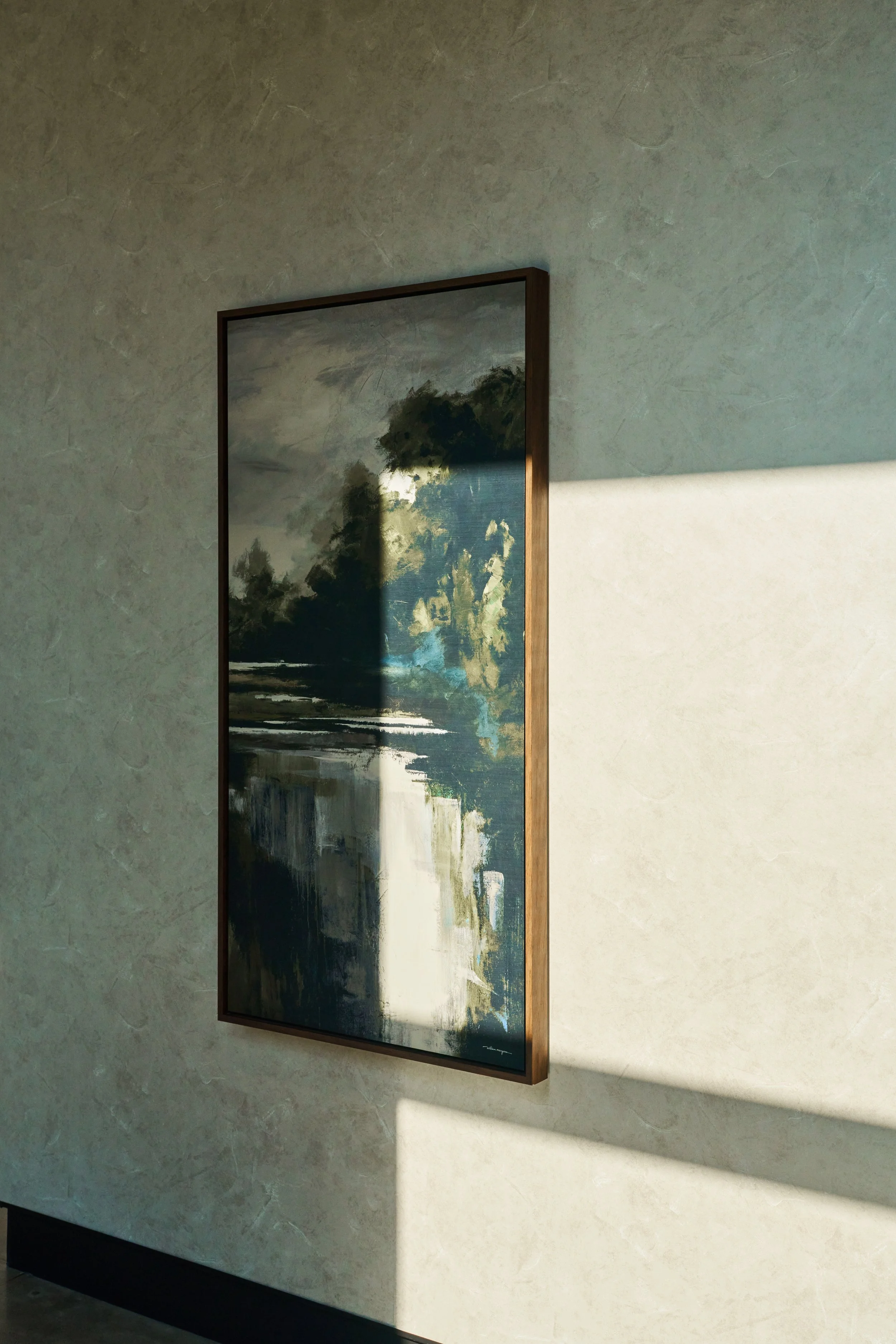Close-up of a landscape painting in a thin wooden frame hanging on a textured beige wall, with sunlight casting a shadow on the wall.