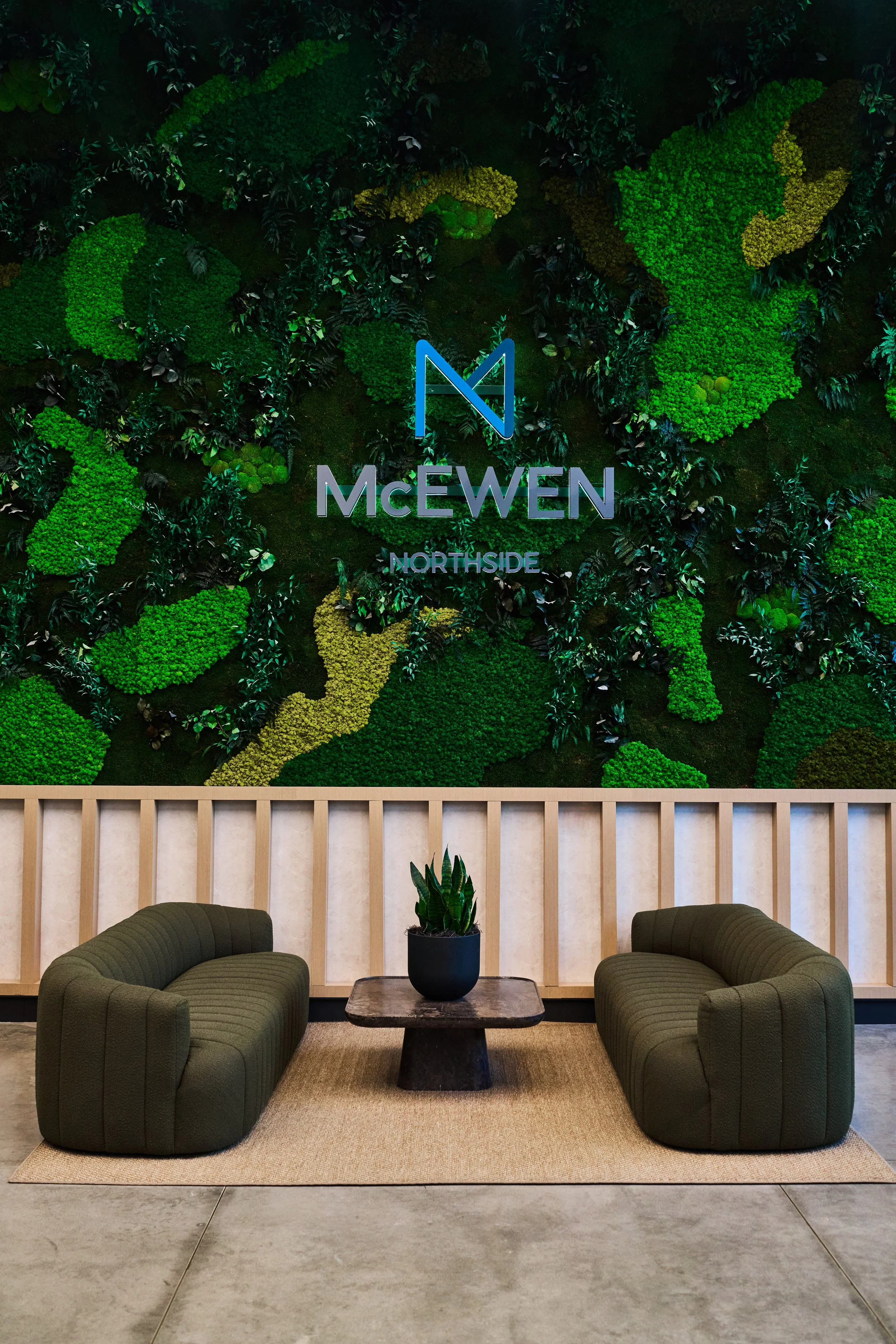 A modern lobby area with two green upholstered armchairs, a dark wooden side table with a potted plant, a beige area rug, a wood-paneled wall, and a large green wall with plants and the logos for McEwen Northside.