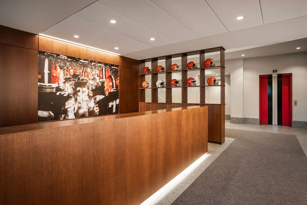 An interior with a wood reception desk, a large framed football-themed black and white photo on the wall, a wooden helmet display shelf with red and orange football helmets, and an elevator with red doors.