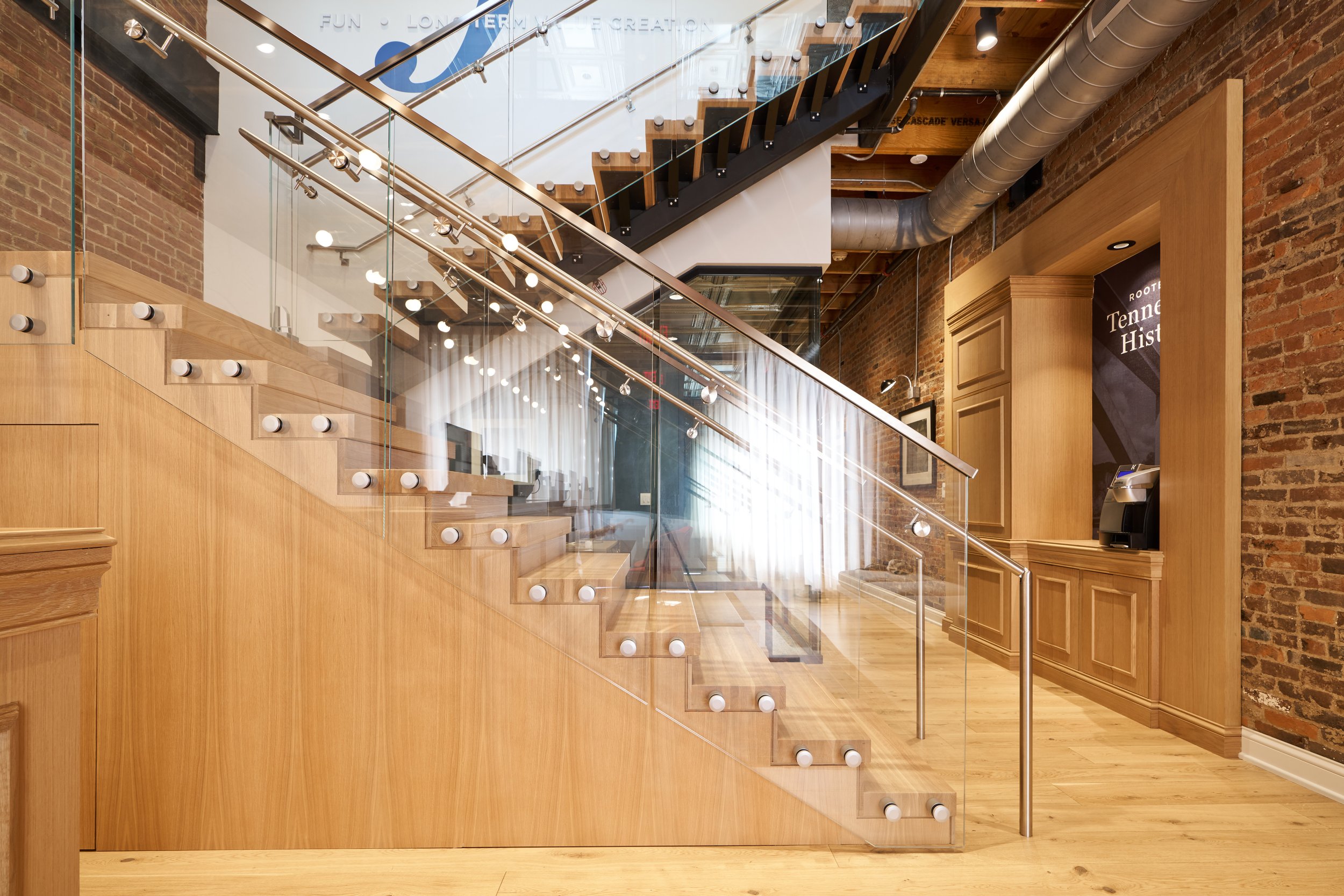 Interior of a modern building with a glass staircase, wooden accents, exposed brick walls, and industrial piping overhead.