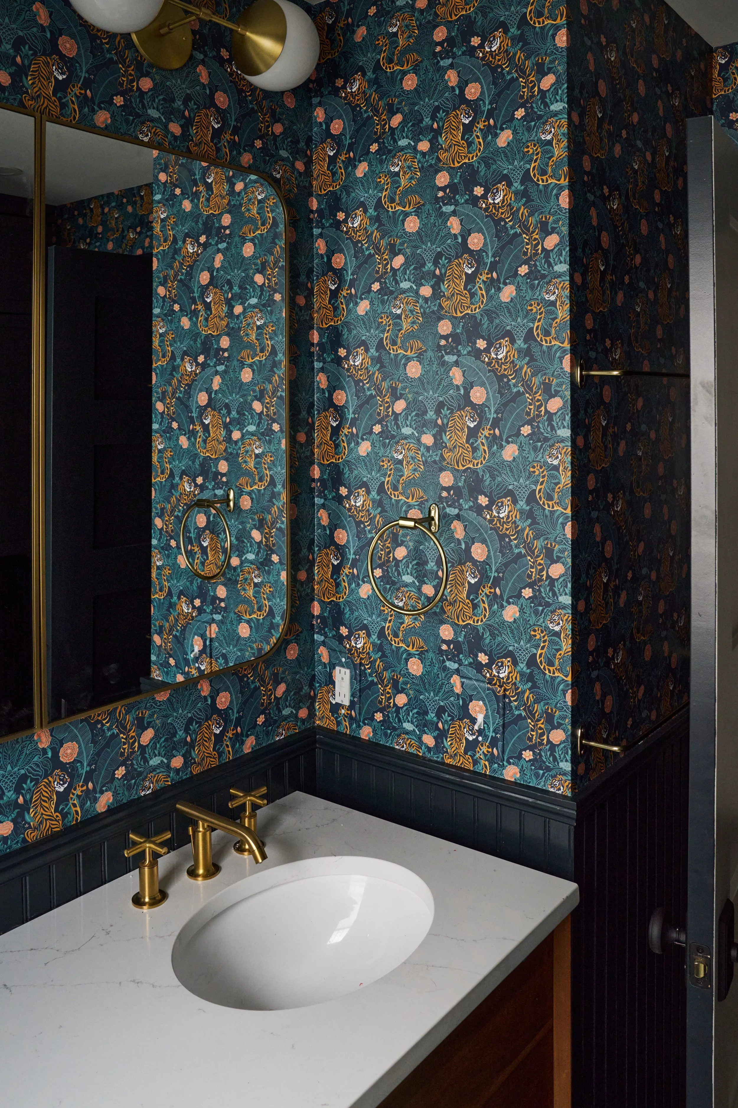 A bathroom with dark floral wallpaper featuring tigers and flowers, a white marble countertop with a round undermount sink, and gold fixtures including faucets and towel bars. There is a mirror with a gold frame reflecting part of the room and a whit