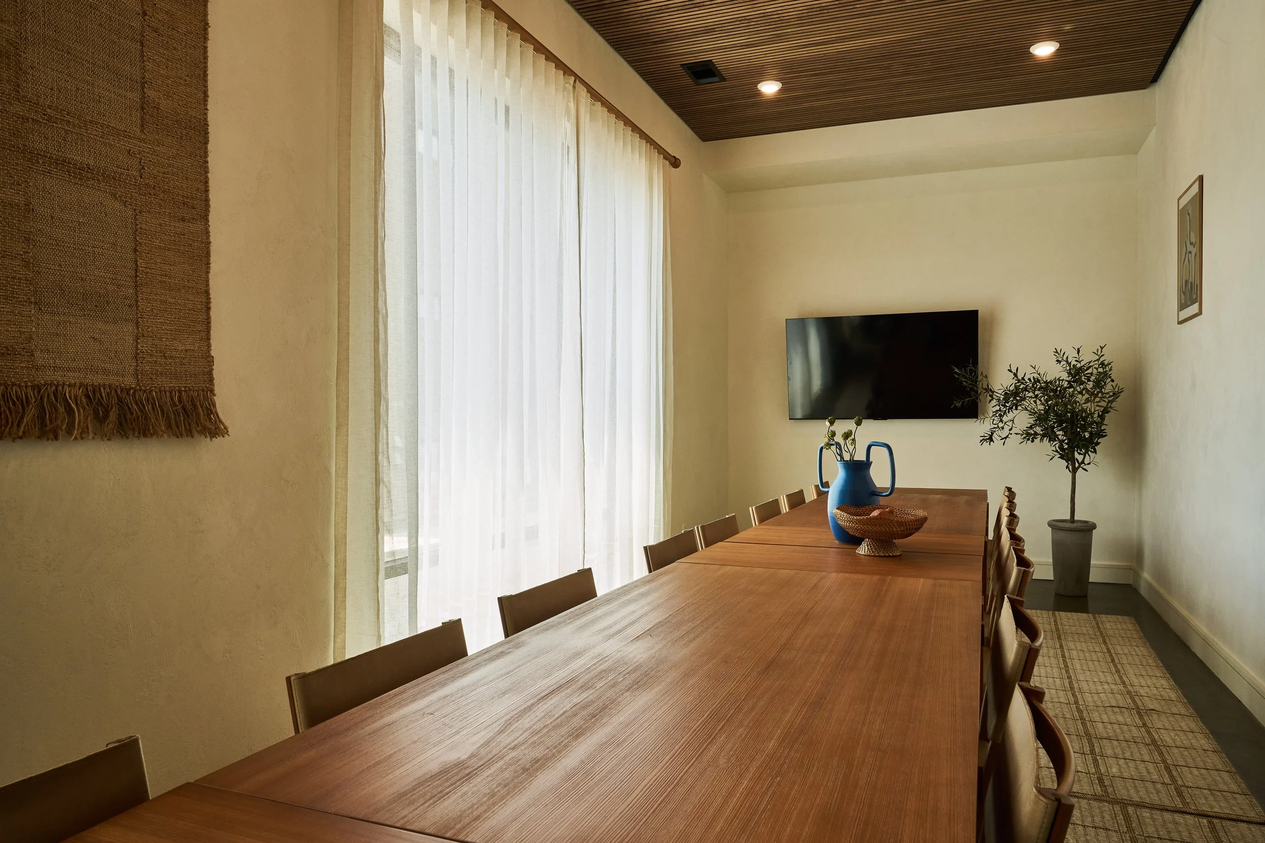 A dining room with a long wooden table surrounded by chairs, decorated with a blue vase holding green plants, and a potted plant in the corner. The room features cream-colored walls, a large window with light curtains, a wall-mounted TV, and a framed