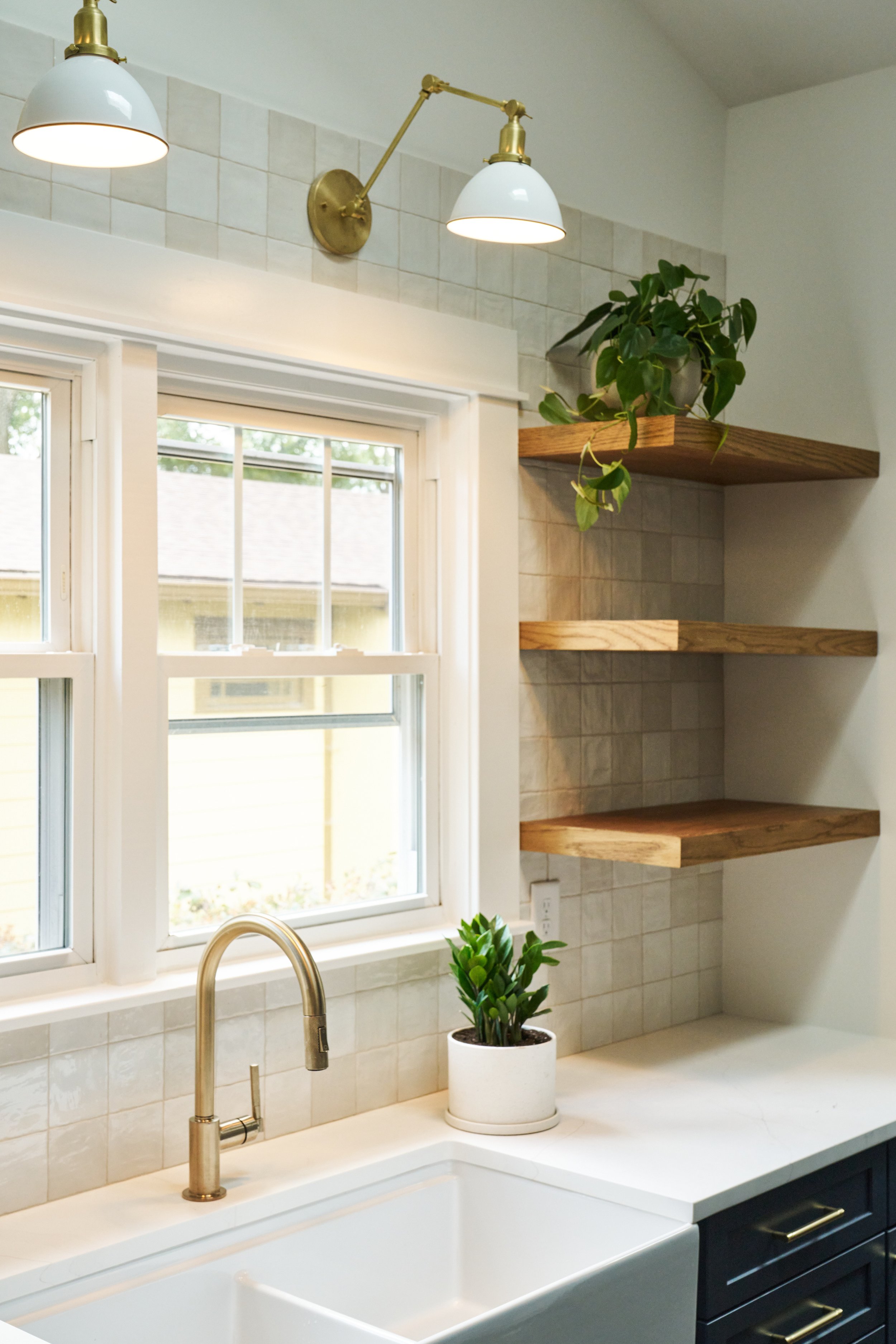 Kitchen with a white sink, a chrome faucet, a potted green plant on the countertop, a window, and three wooden floating shelves with a plant, fitted with brass lighting fixtures.