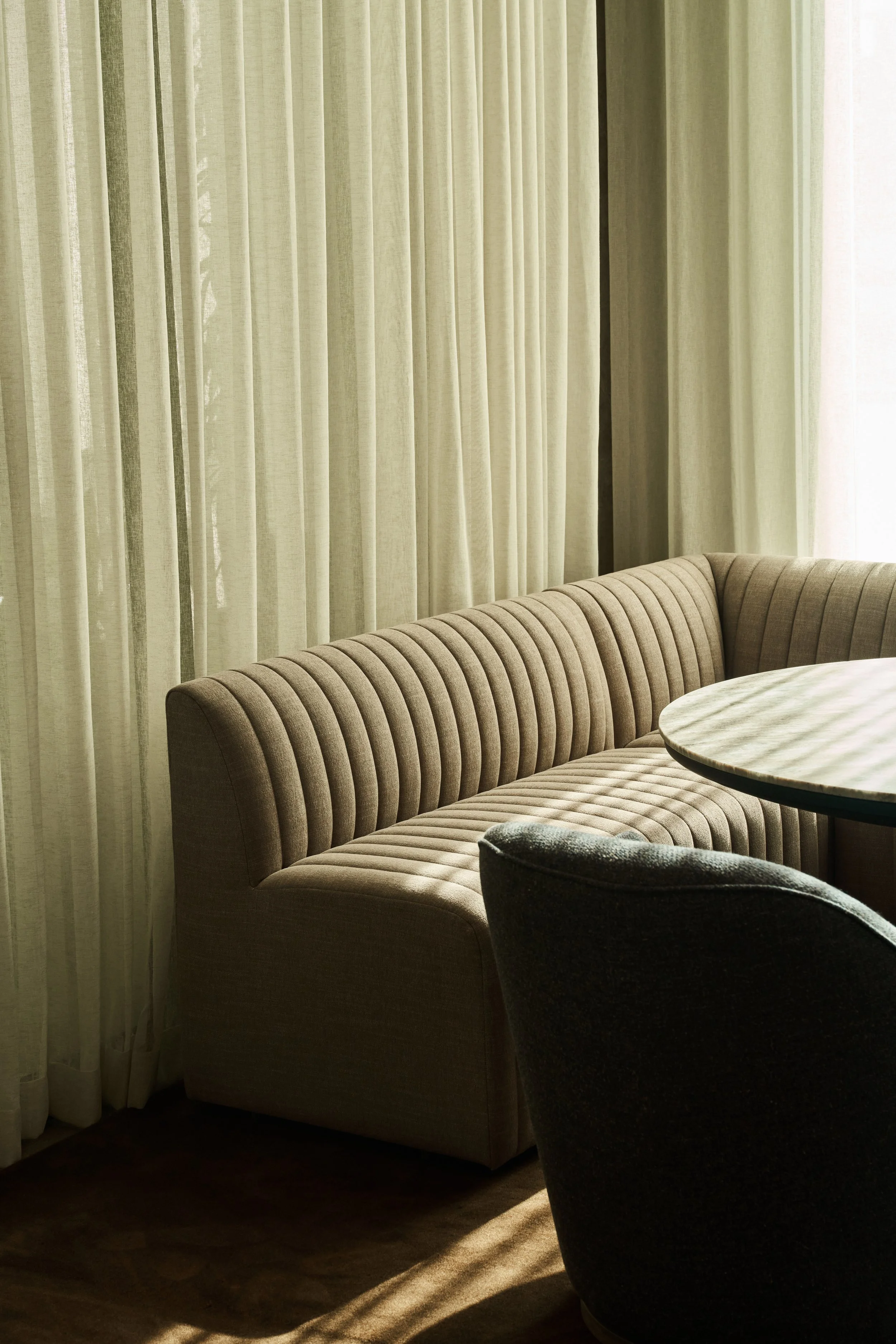 Room with beige striped upholstered sofa, dark upholstered armchair, and light-colored curtains covering large windows, illuminated by natural sunlight.