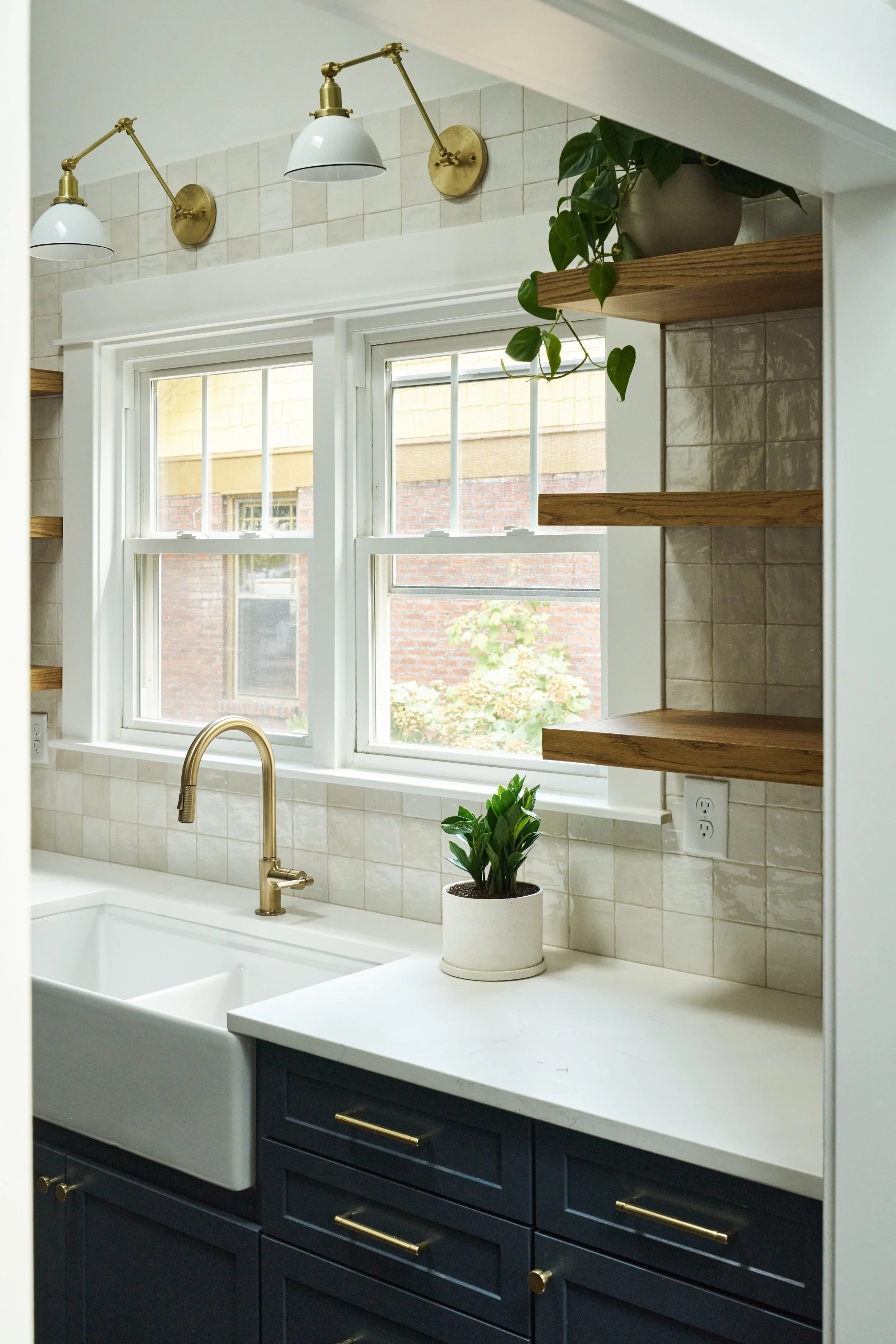 A kitchen window with white trim, a white sink, a gold faucet, a potted plant on a white countertop, blue kitchen cabinets with gold hardware, wooden floating shelves, and tile backsplash. Two adjustable white and gold wall lamps are near the window.