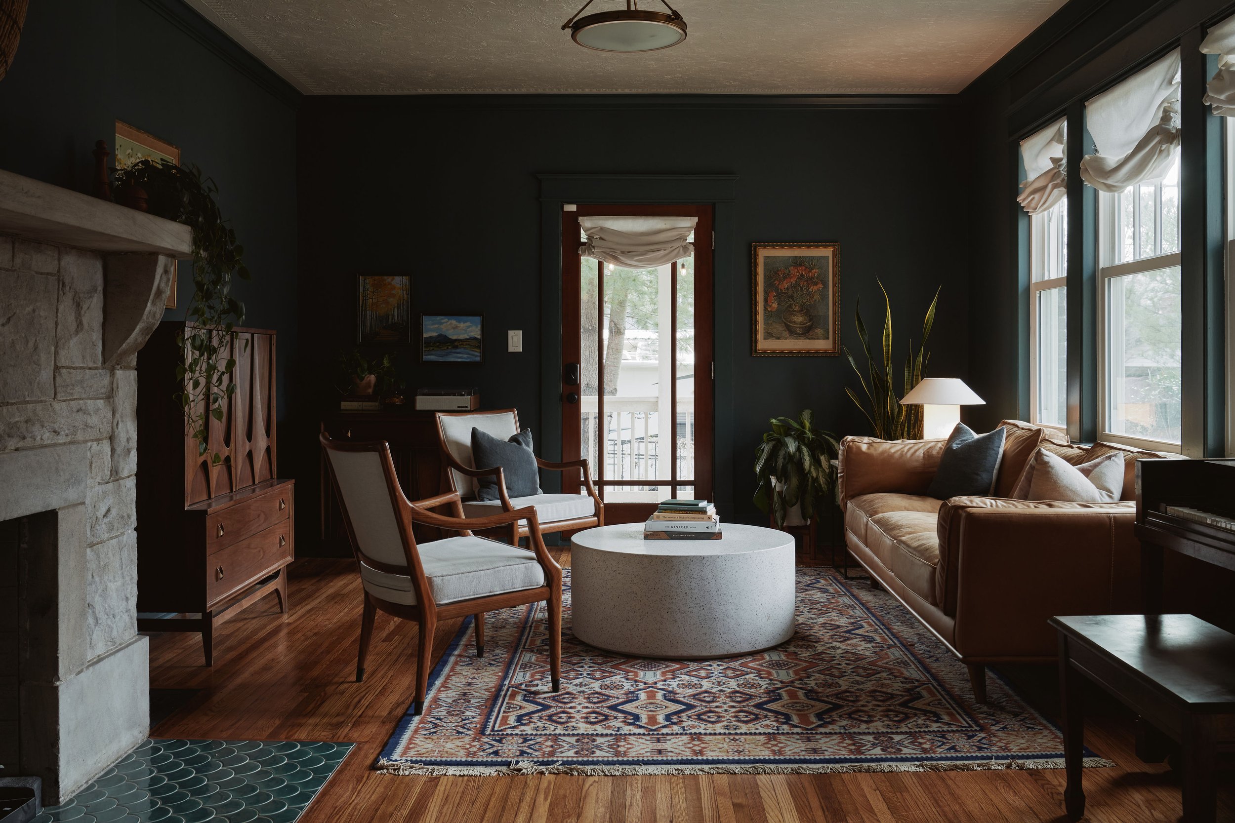 Living room with dark walls, wooden chairs, a sofa, a stone fireplace, framed artwork, a window with curtains, a rug, a side table with books, and plants.