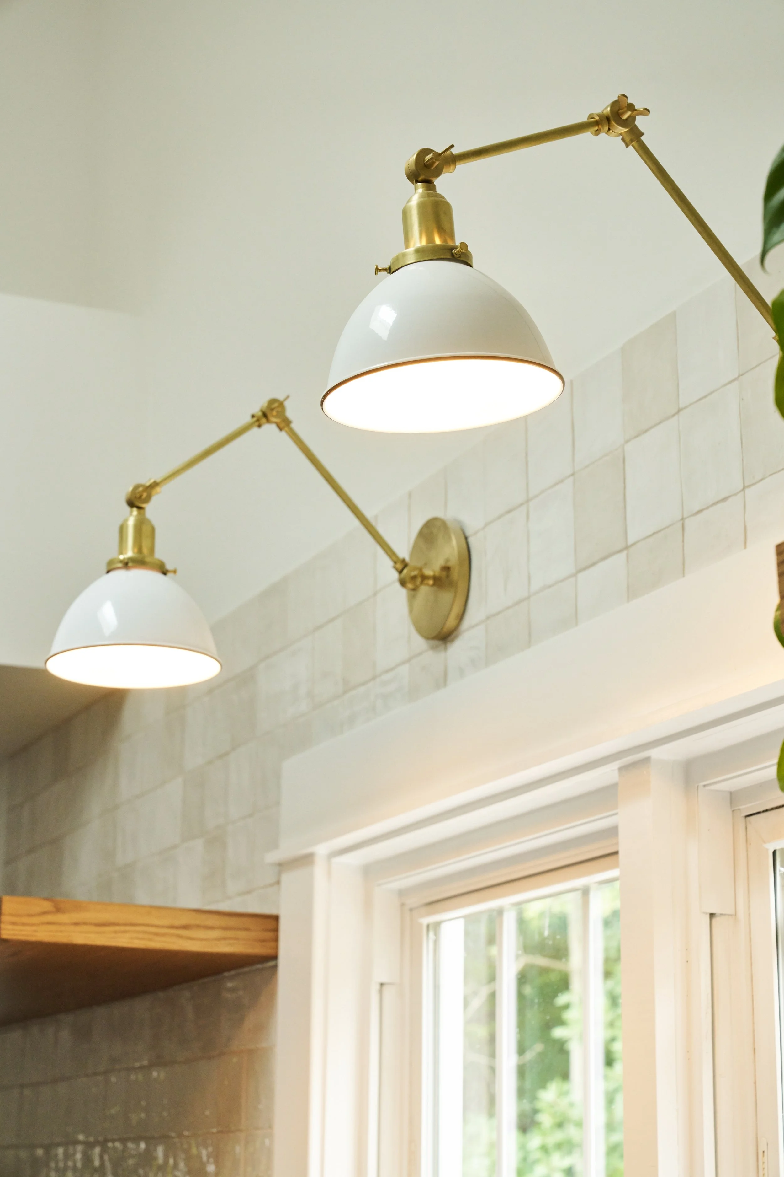 Close-up of two wall-mounted white lamps with brass arms, illuminating a tiled wall near a window in a room with natural light.