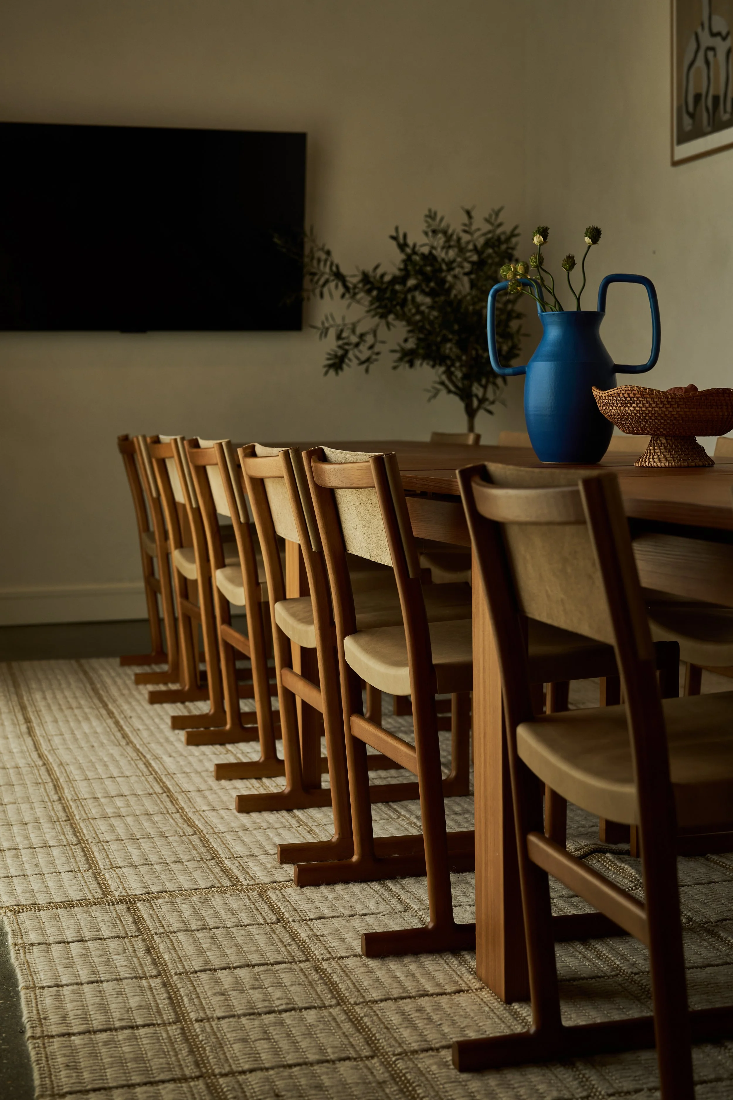 A long wooden dining table with beige chairs, a blue vase with green flowers, a woven basket, and a plant in the background in a room with beige walls and a patterned rug.