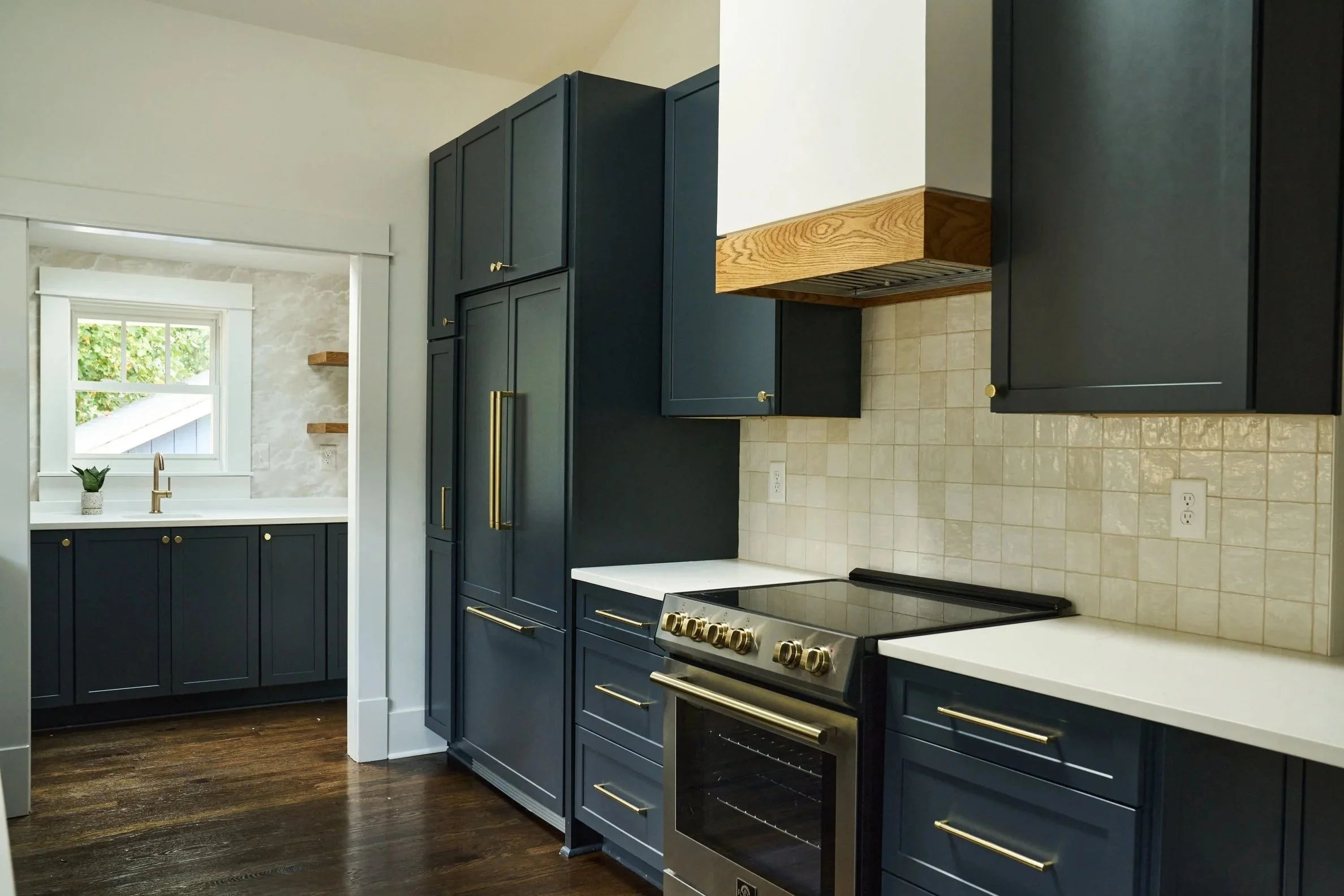 View of a modern kitchen with dark blue cabinets, a stove, countertops, a window with a small plant, and beige tiled backsplash.