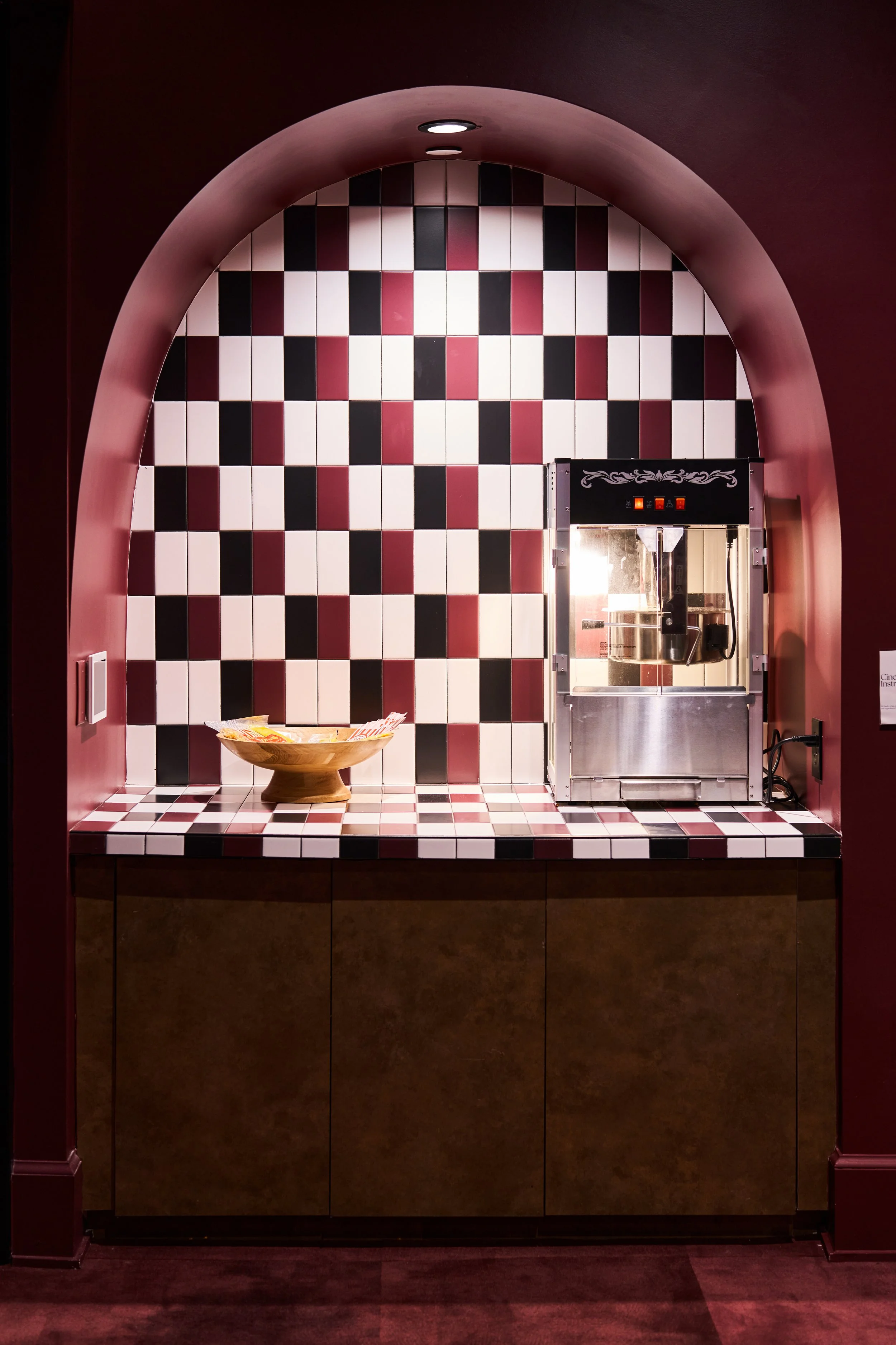 A tiled wall in black, white, and red checkered pattern with an arched opening framing the scene. Inside, there is a popcorn machine and a decorative bowl with snacks on a matching tiled counter, all set against a dark background.