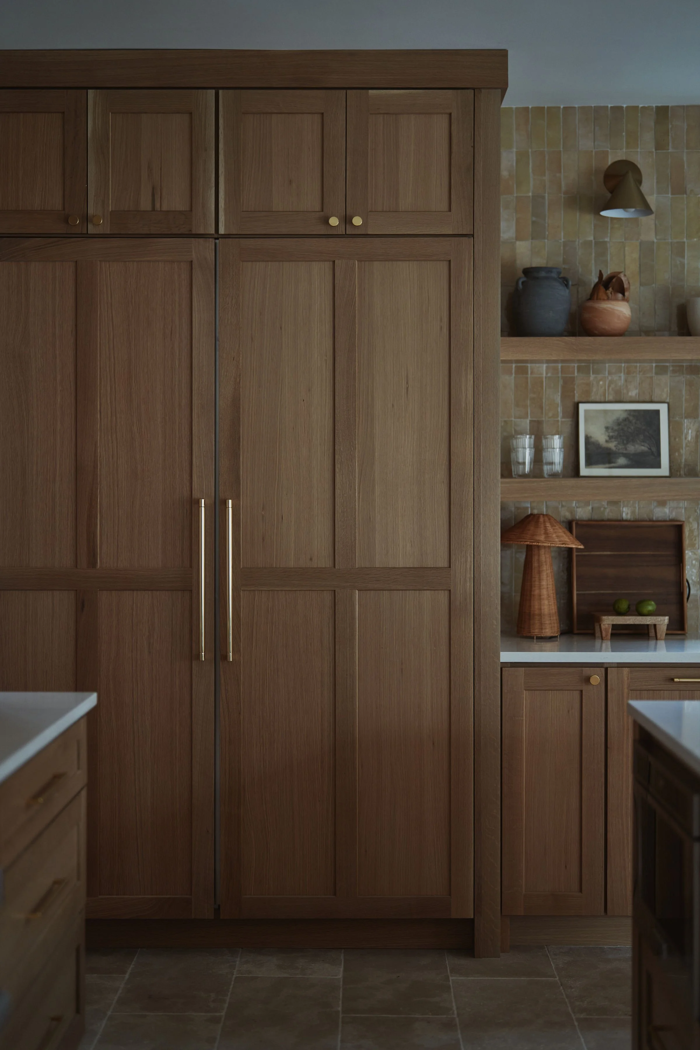 A wooden kitchen cabinet with a natural wood finish and two vertical brass handles in front of a beige tiled wall. To the right, decorative pottery, a framed photo, and a lamp are visible on the shelves.