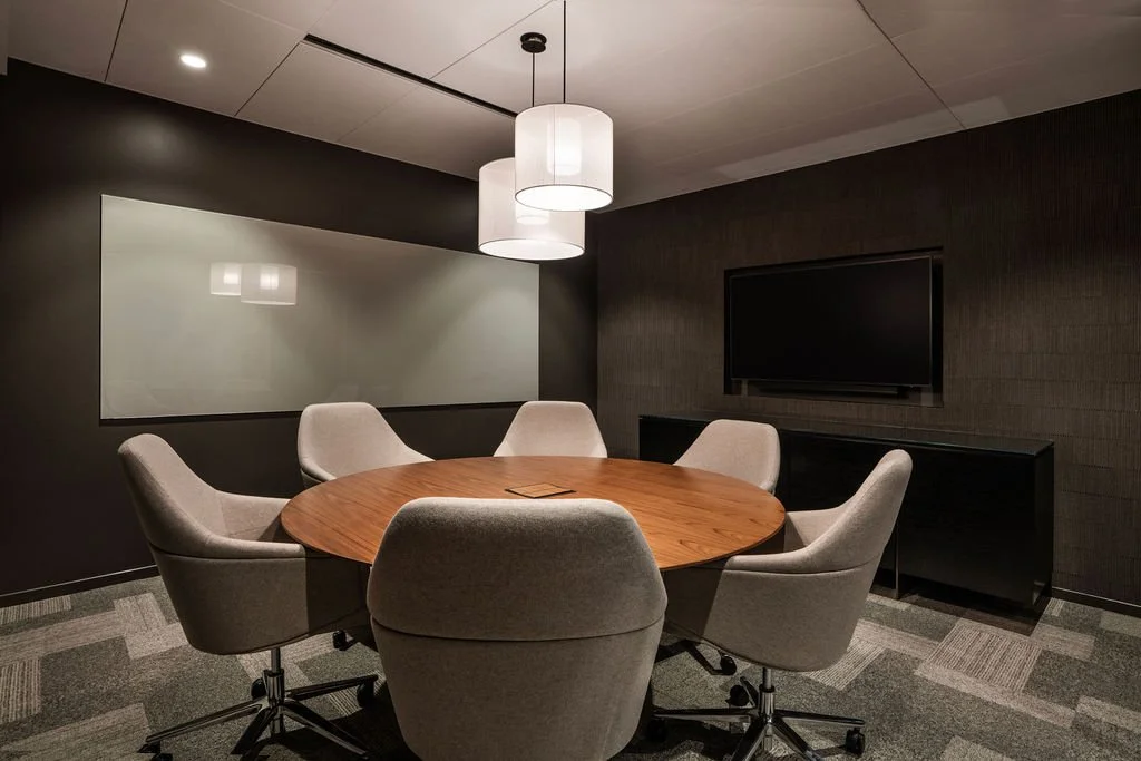 A modern conference room with a round wooden table surrounded by six light-colored upholstered chairs. There is a large flat-screen TV on the dark textured wall, a black cabinet beneath it, and a whiteboard on the wall opposite the TV. Two pendant li