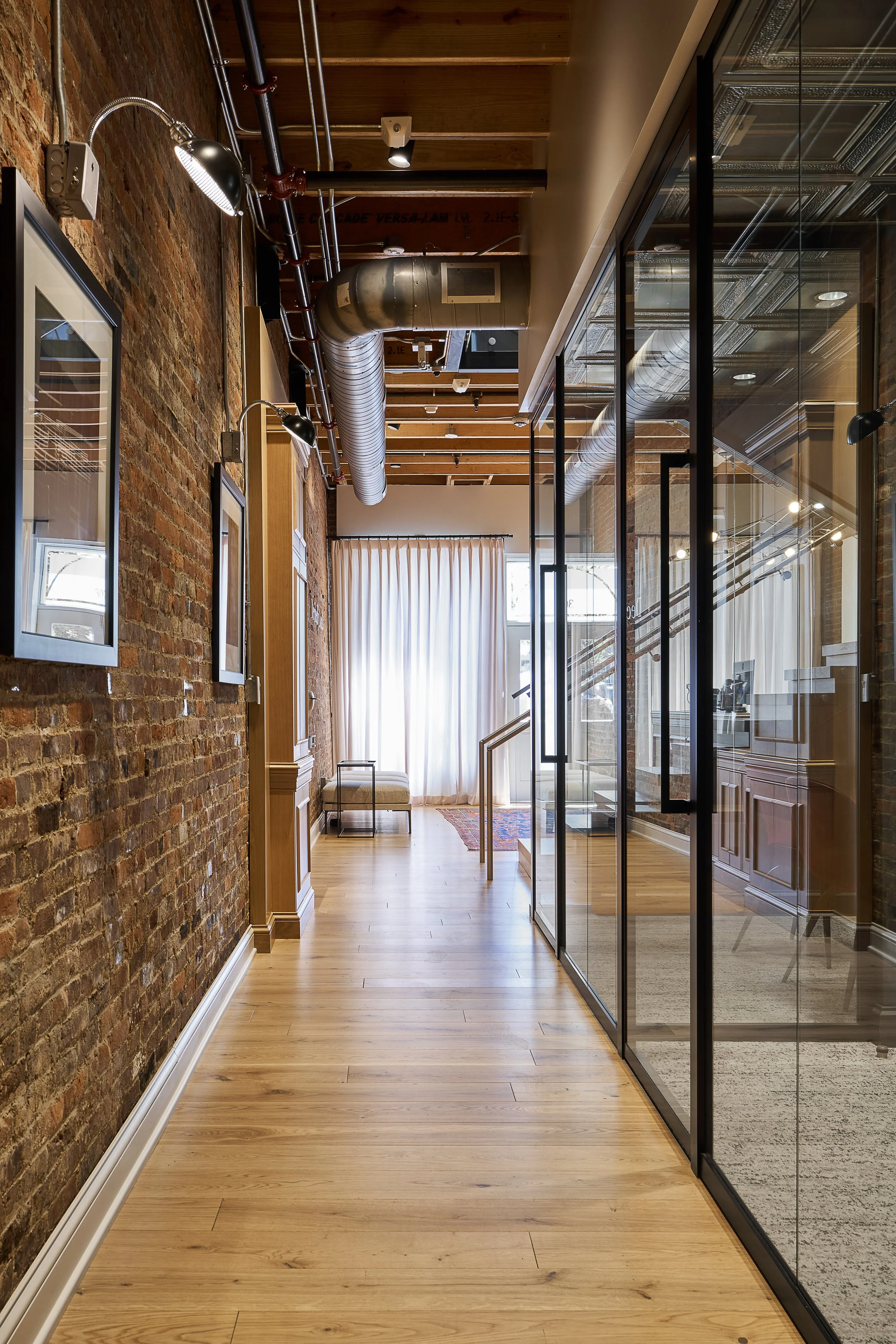 A modern interior hallway with exposed brick walls, wooden flooring, glass partitions with black frames, and ceiling with air ducts and lighting fixtures. At the end, there are curtains and a small bench.