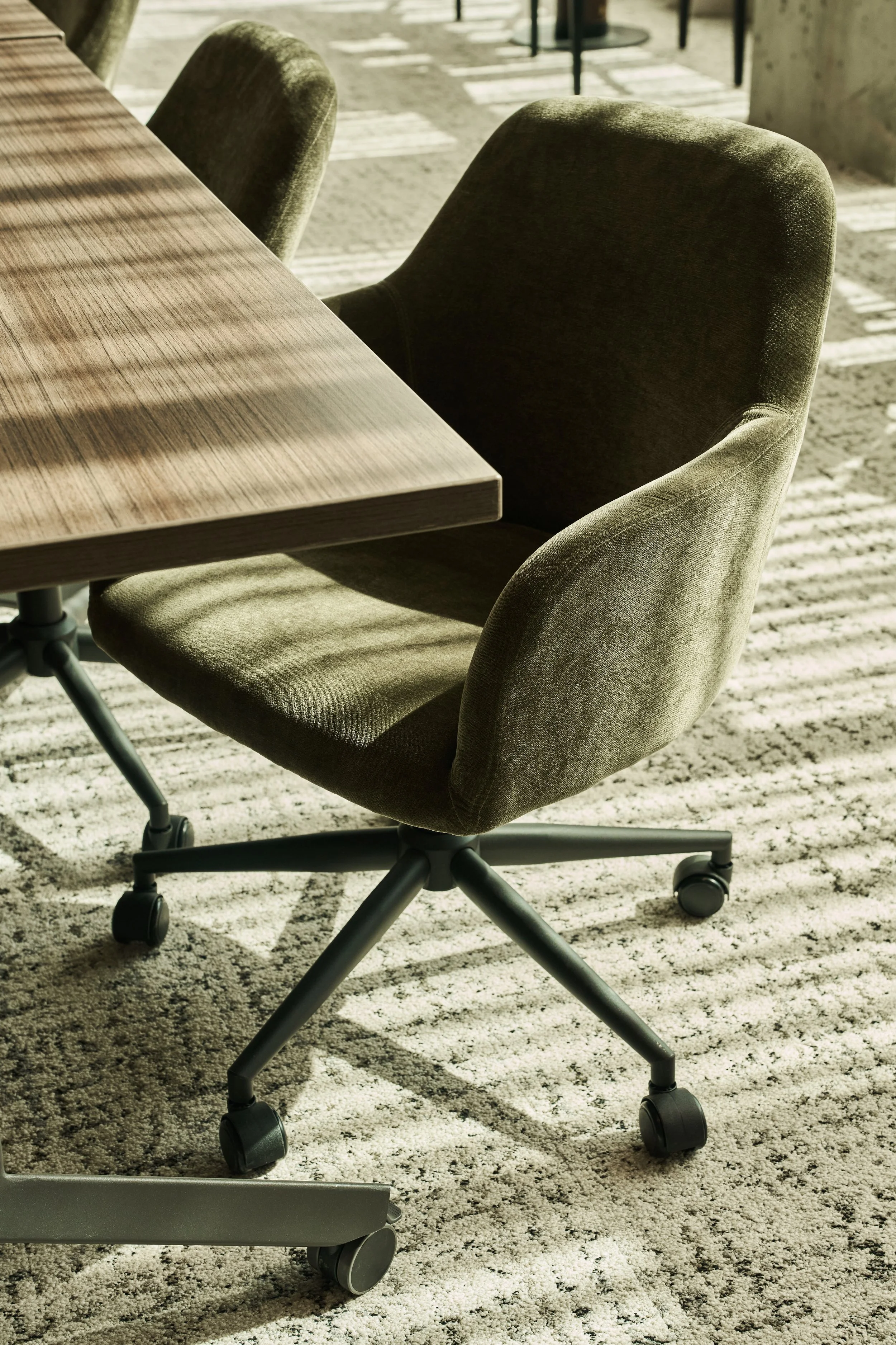 Office chair with a rounded backrest and black wheels on a beige and black patterned carpet, next to a wooden meeting table with a metal column base, in a bright room with sunlight and shadows.