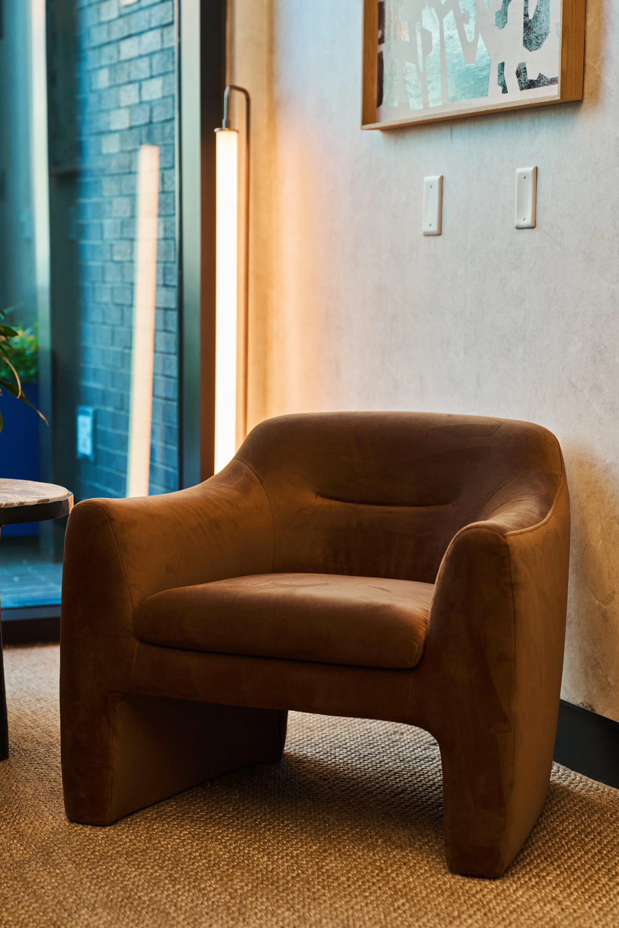 A cozy corner with a brown leather armchair, a small side table, and a textured beige carpet. There is a doorway with a glass door and brick wall on the left, a wall with a framed abstract artwork above the armchair, and a modern vertical light fixtu