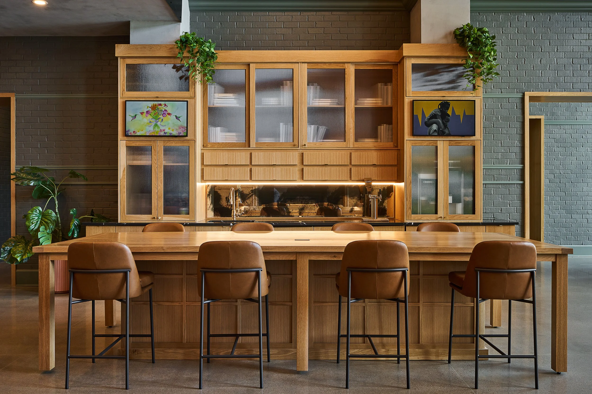 A modern kitchen with a large wooden dining table and six brown leather chairs. Behind the table is a wooden cabinet with glass doors, containing books. The cabinet has plants hanging from its top corners, and two framed artworks on either side. The 