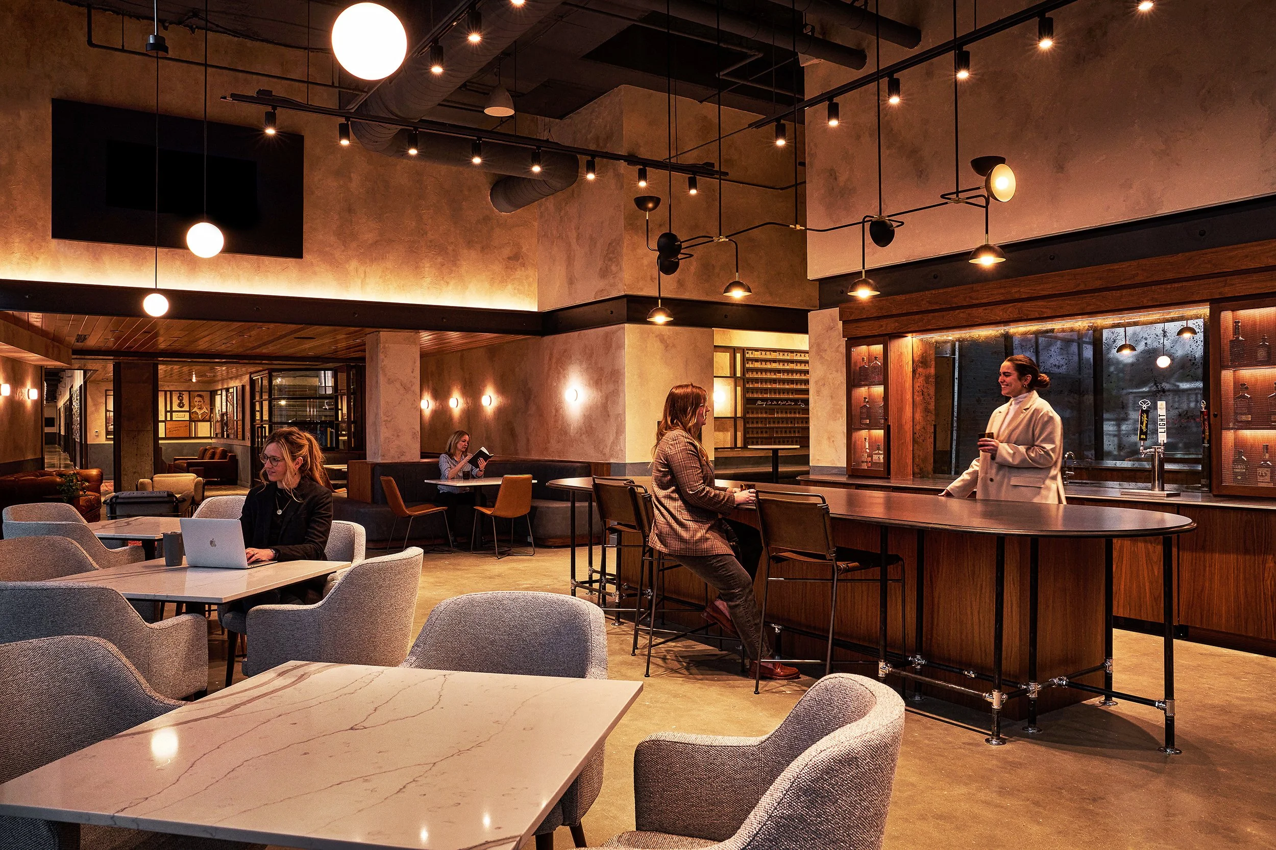 Interior of a modern café or restaurant with warm lighting, wooden accents, and several people seated at tables. One woman is working on a laptop, and others are engaged in conversation or reading. The space has a cozy, inviting atmosphere with dim lighting and contemporary decor.