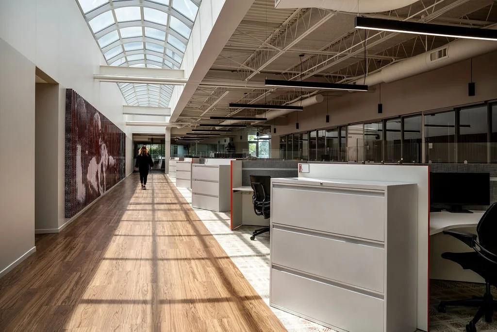Modern office corridor with wooden flooring, white desks, and glass wall partitions, illuminated by natural light from a curved glass ceiling.