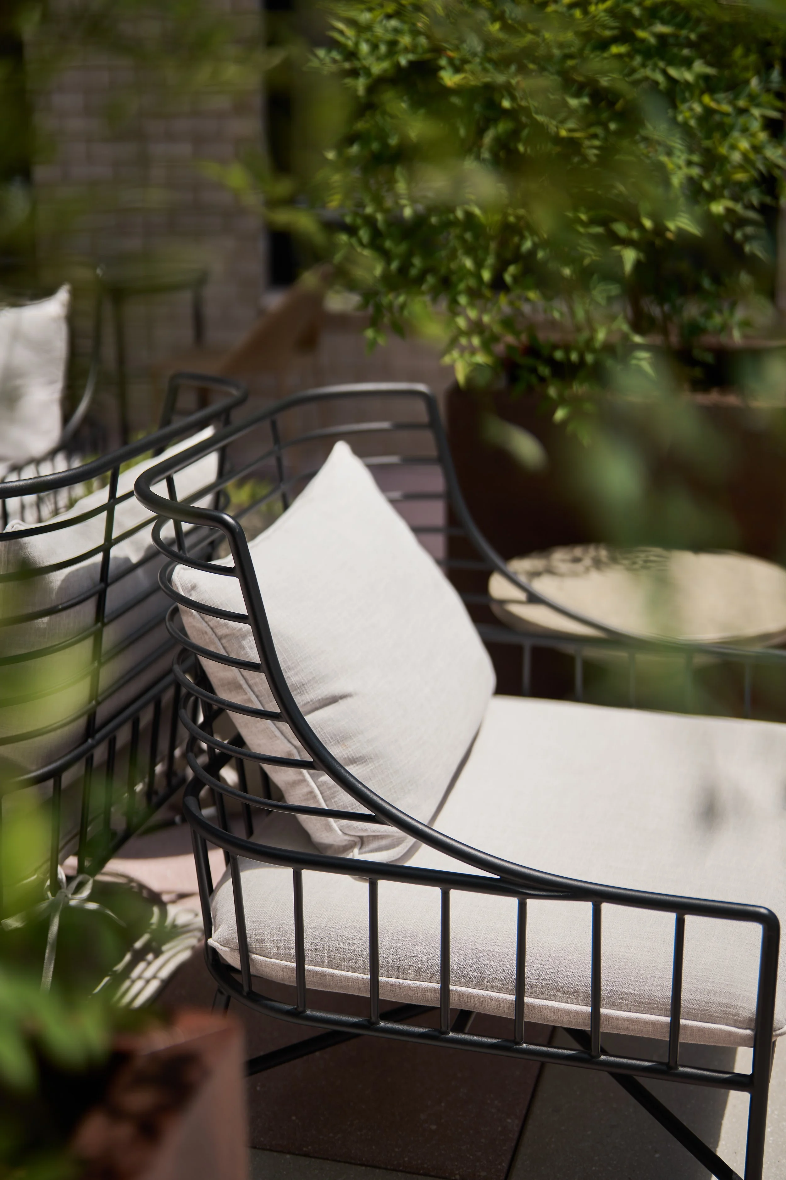 Outdoor patio furniture with metal chairs and beige cushions surrounded by green plants and a brick wall in the background.