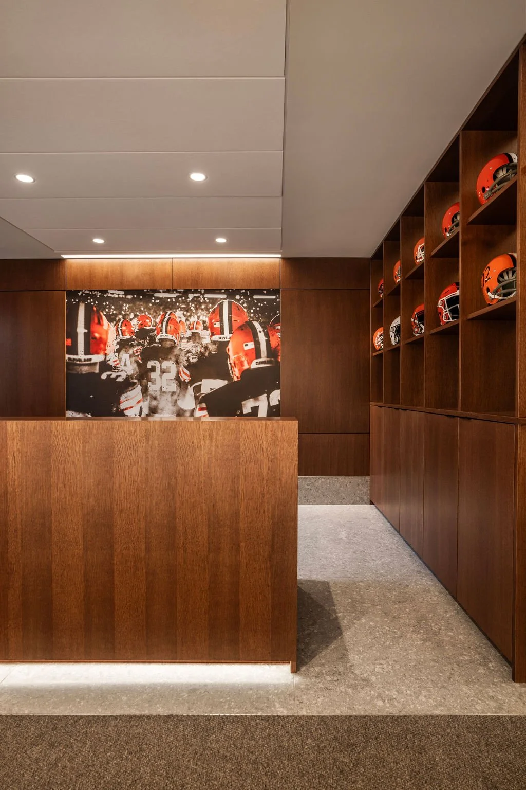 A modern locker room with wooden lockers and shelves, displaying orange football helmets. There is a large black and white photo of football players with helmets on the back wall.