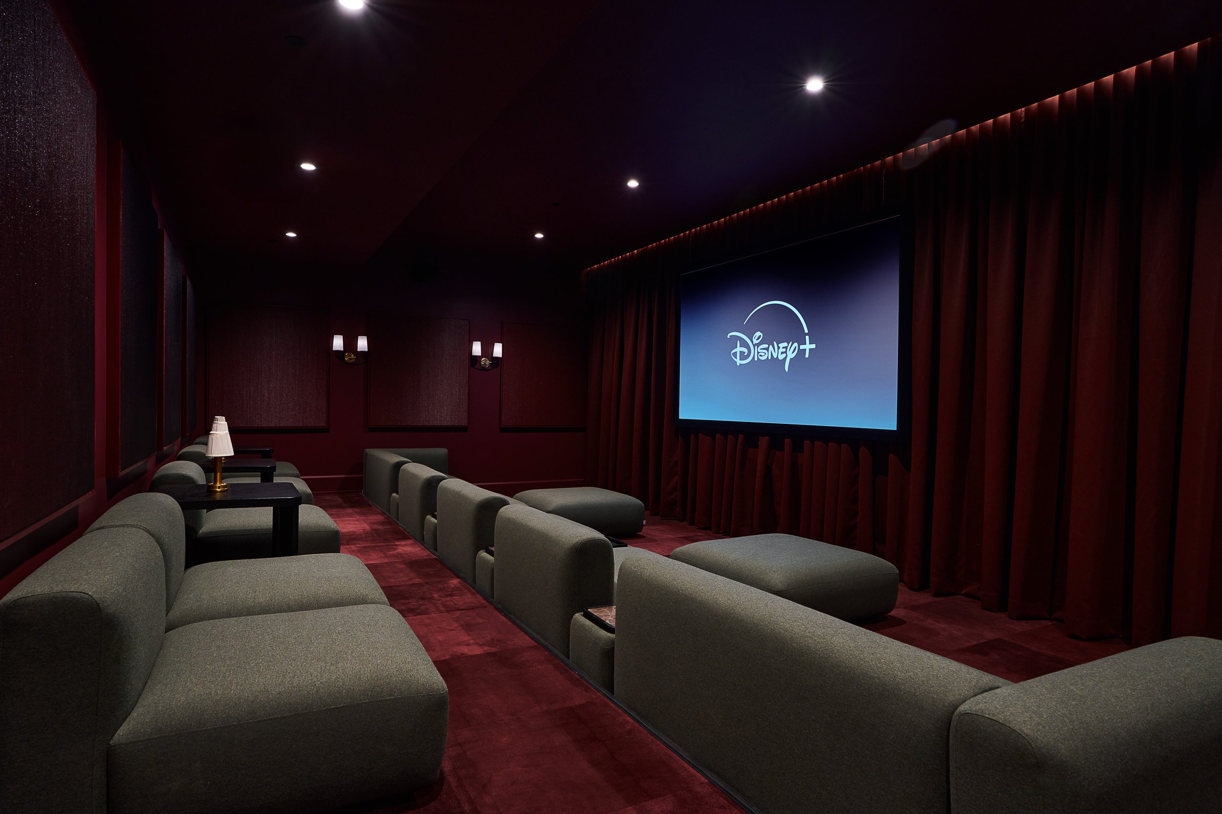 An indoor movie theater room with dark red walls, gray seating, tables with lamps, and a large screen displaying the Disney+ logo.