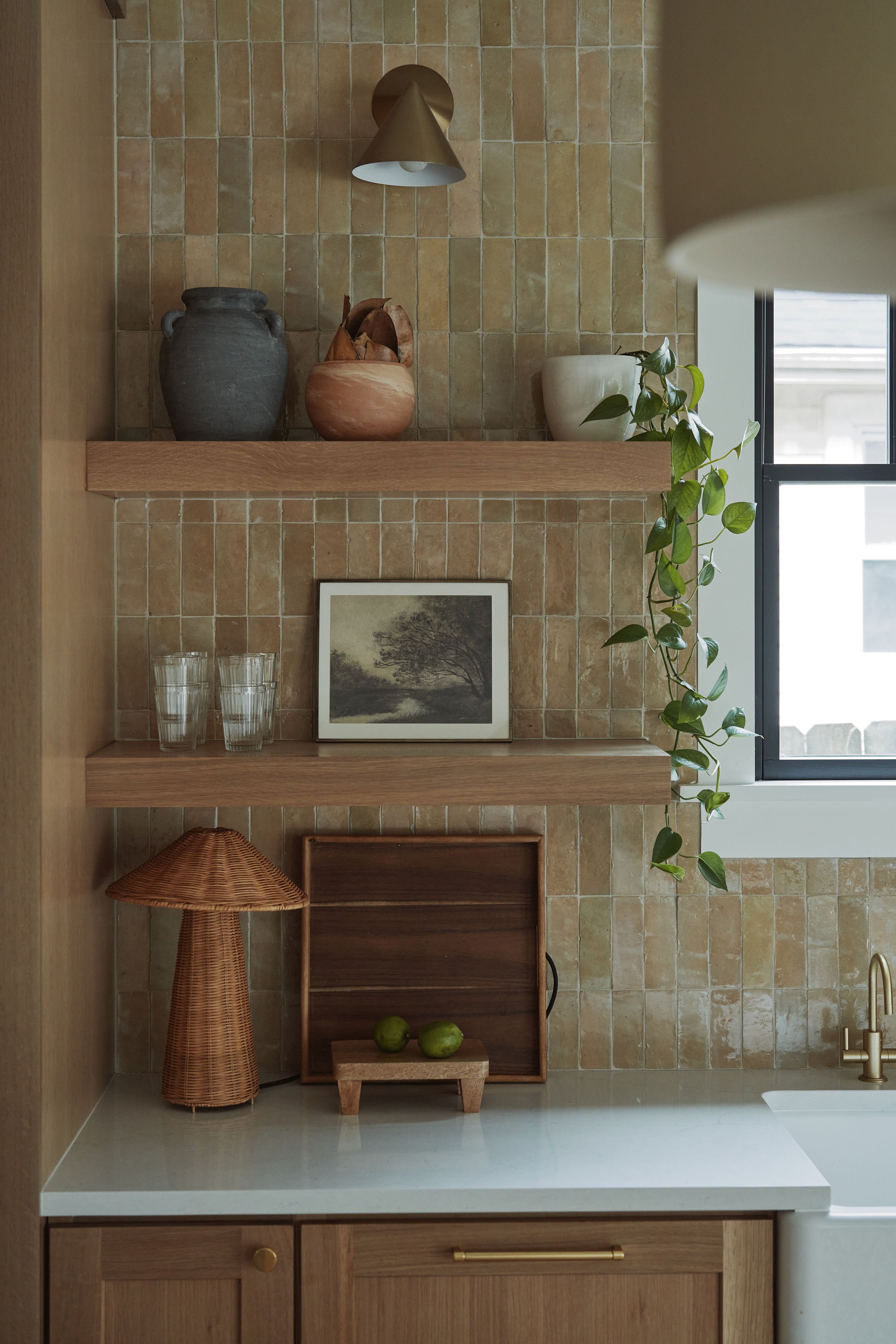 Interior of a kitchen with beige tiled wall, wooden cabinets, and decorative elements including vases, a plant, a framed picture, and a woven lamp.