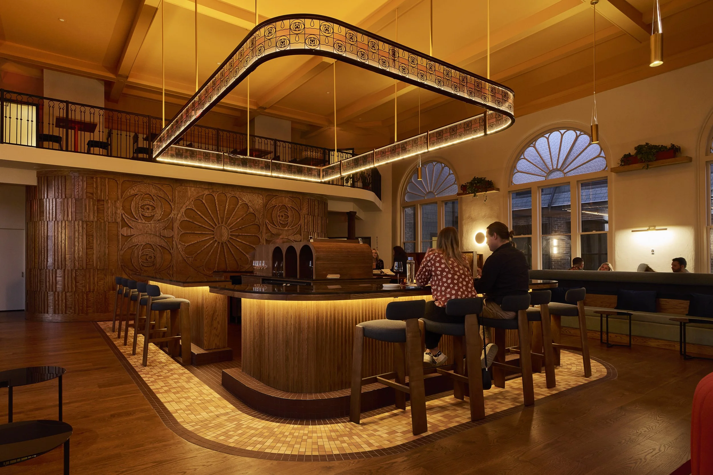 Inside a warmly lit restaurant with a high ceiling, large arched windows, and a wooden spiral staircase. There are people seated at a bar with wooden accents and a high ceiling, with some patrons engaged in conversation.