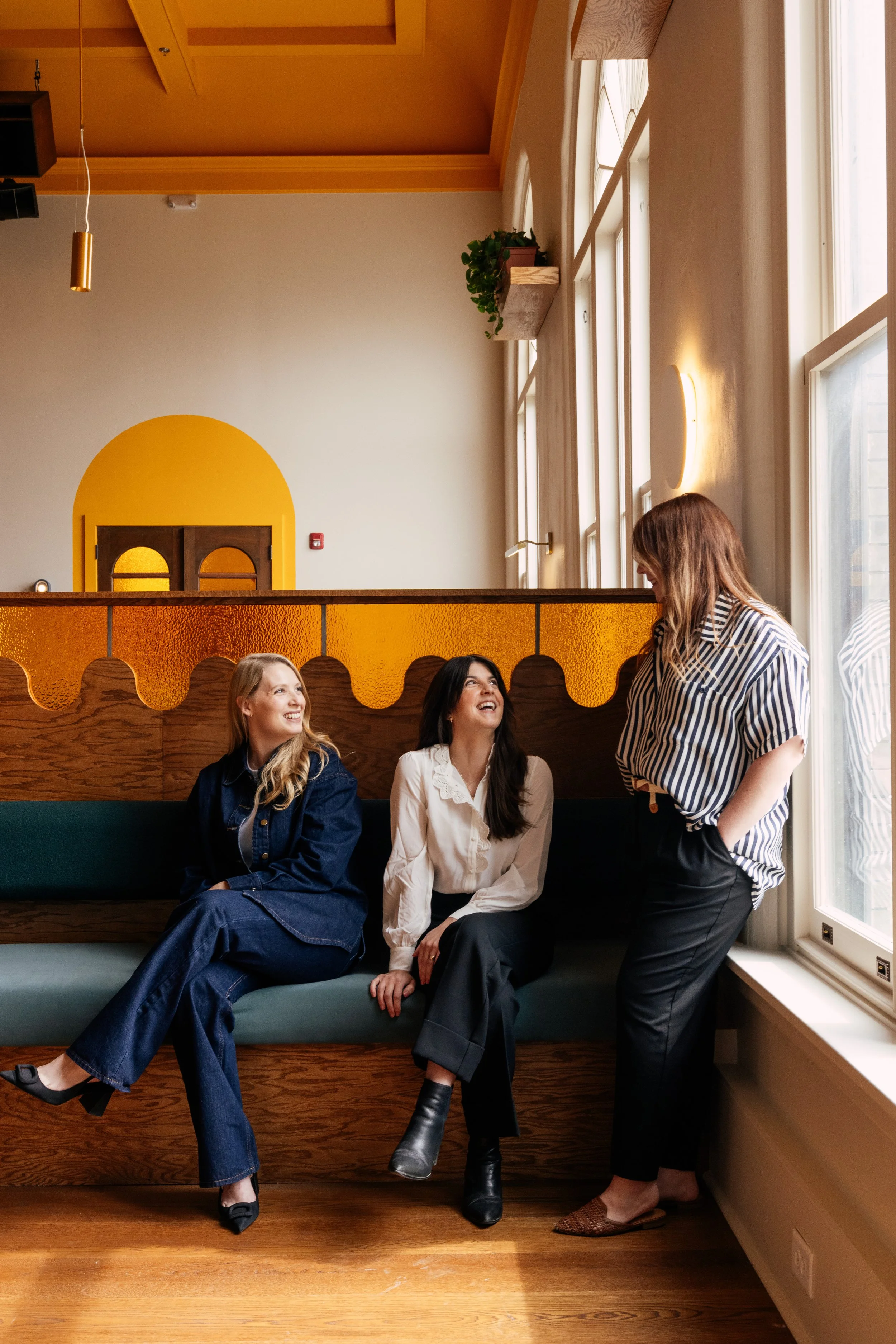 Four women engaging in a conversation inside a stylish café with wooden floors, a teal cushioned bench, and large windows letting in natural light; they are smiling and dressed in casual, fashionable clothing.