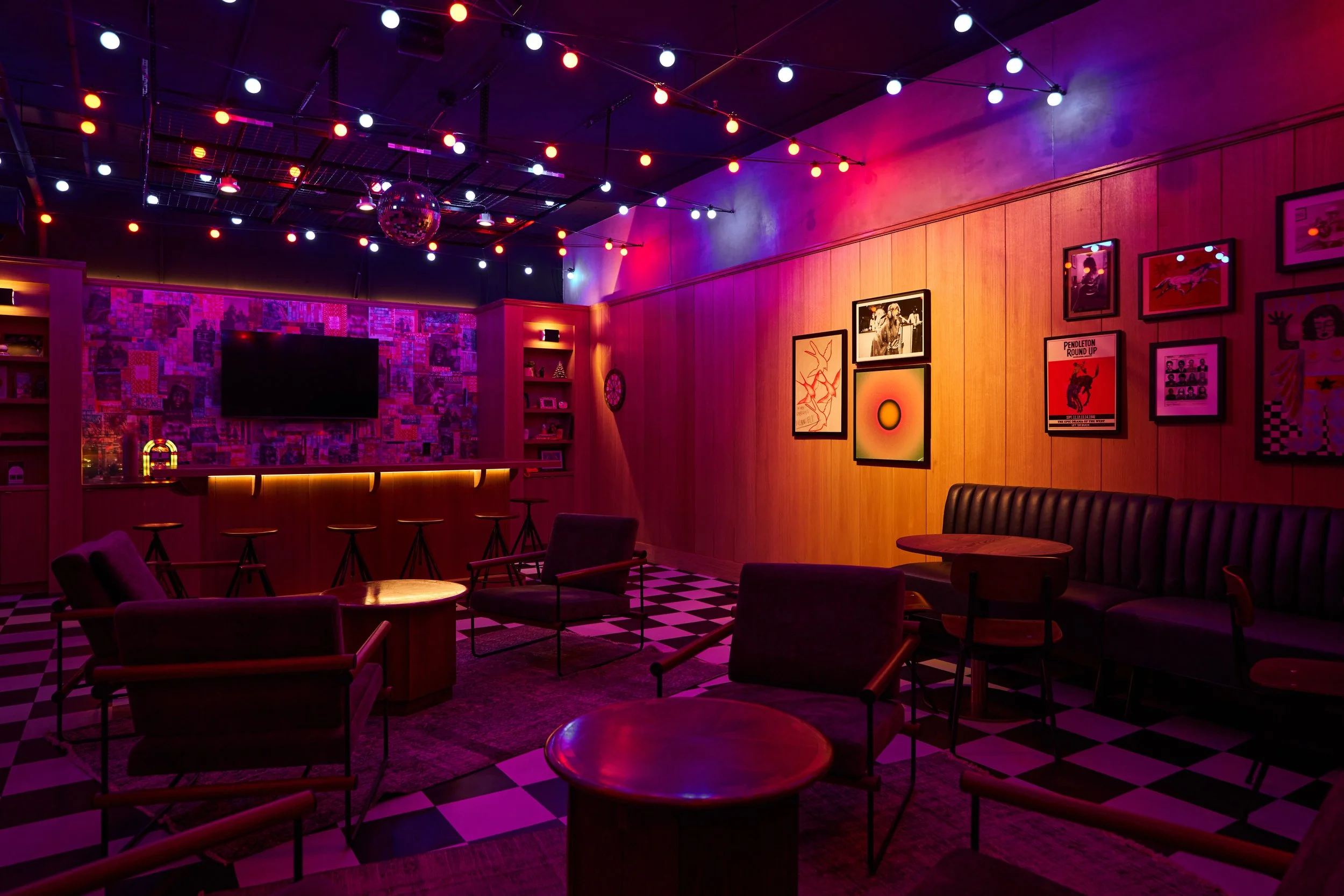 A dimly lit lounge with purple, pink, and red lighting, decorated with framed artwork and photos, with a checkered black and white floor, and scattered tables and chairs.