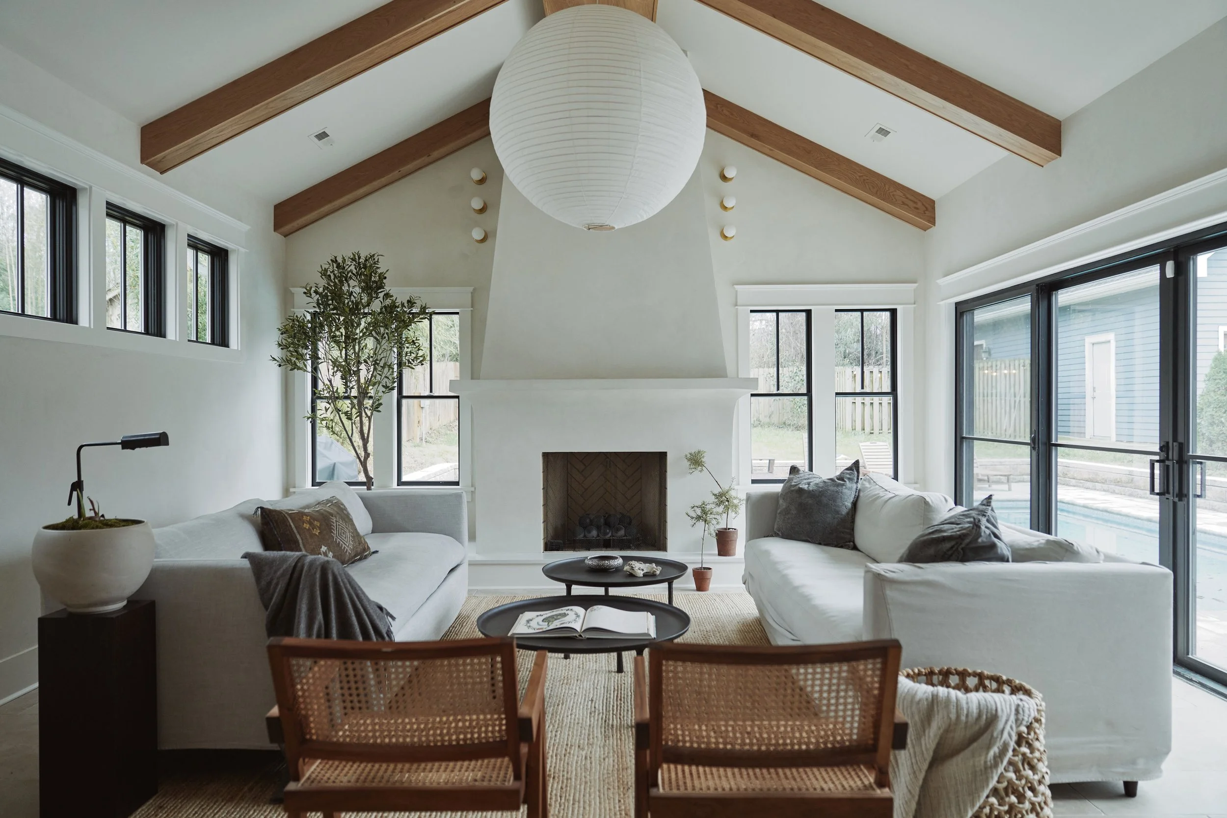 Living room with high ceiling, wooden beams, large white paper lantern, white sofas, potted plants, fireplace, and large windows and glass doors allowing outdoor view.