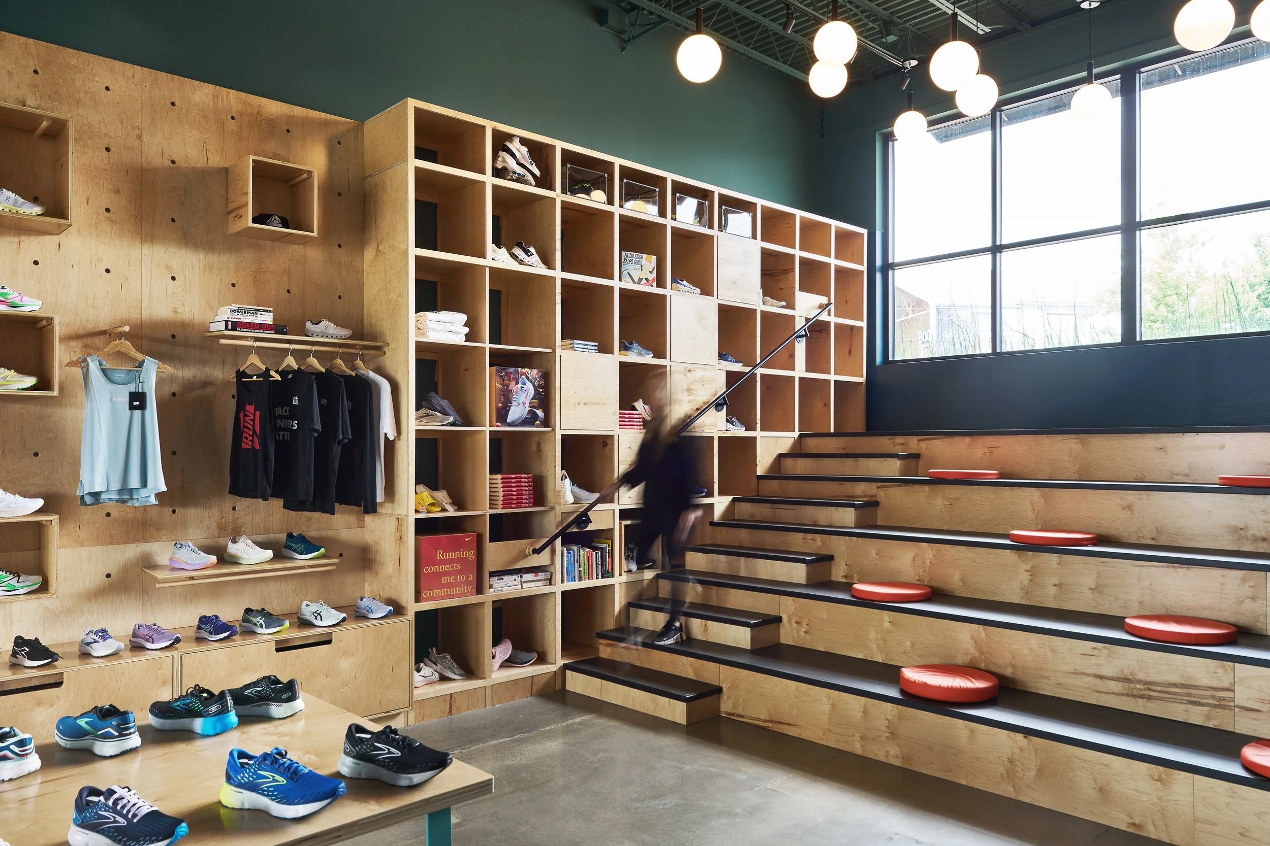 Interior of a retail store displaying athletic shoes, clothing, and books on wooden shelves, with large windows, modern lighting, and seating stairs.