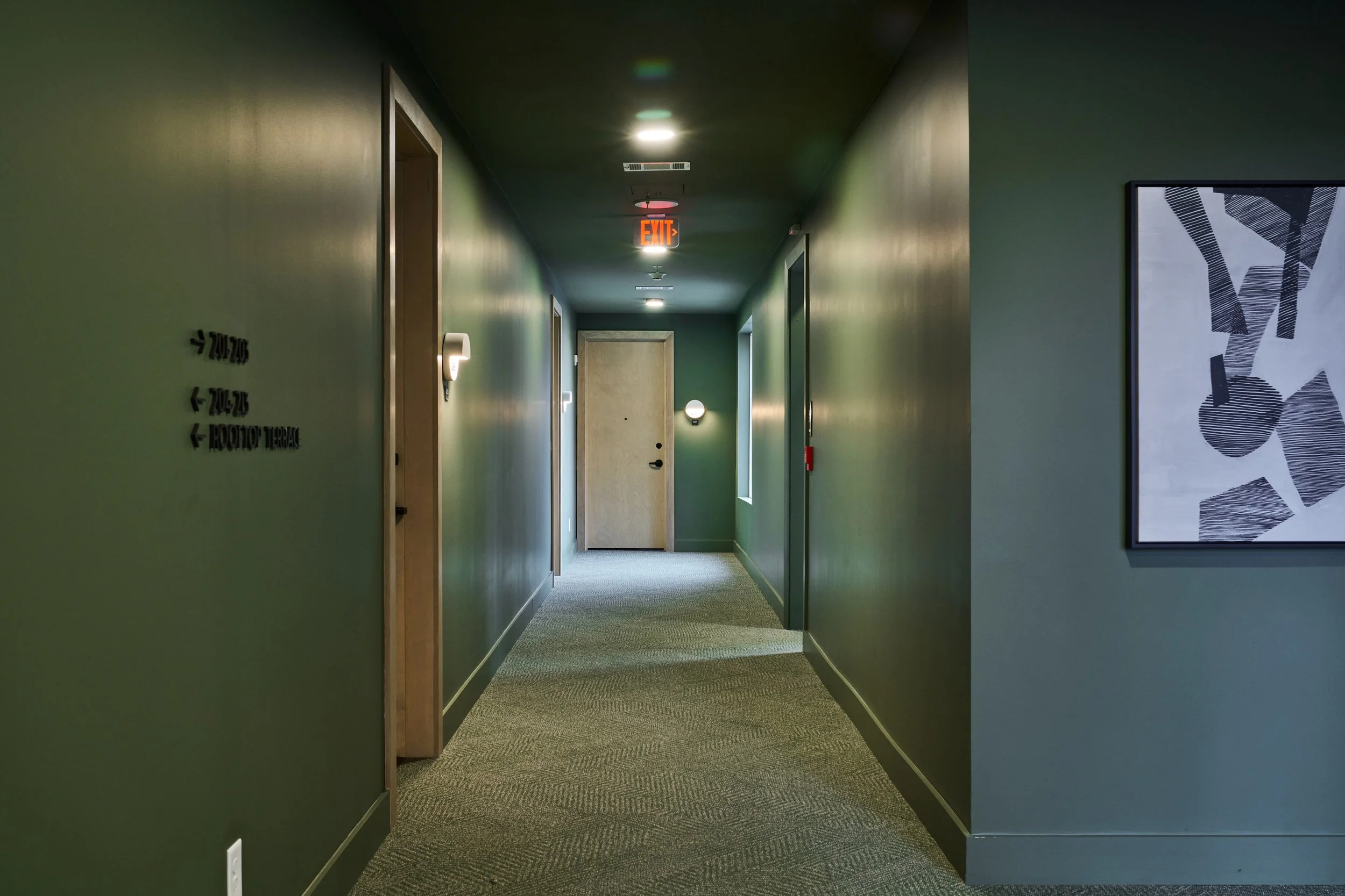 Long indoor corridor with green walls, illuminated by ceiling lights and wall-mounted lamps, leading to a closed door at the end, with an illuminated exit sign hanging from the ceiling.