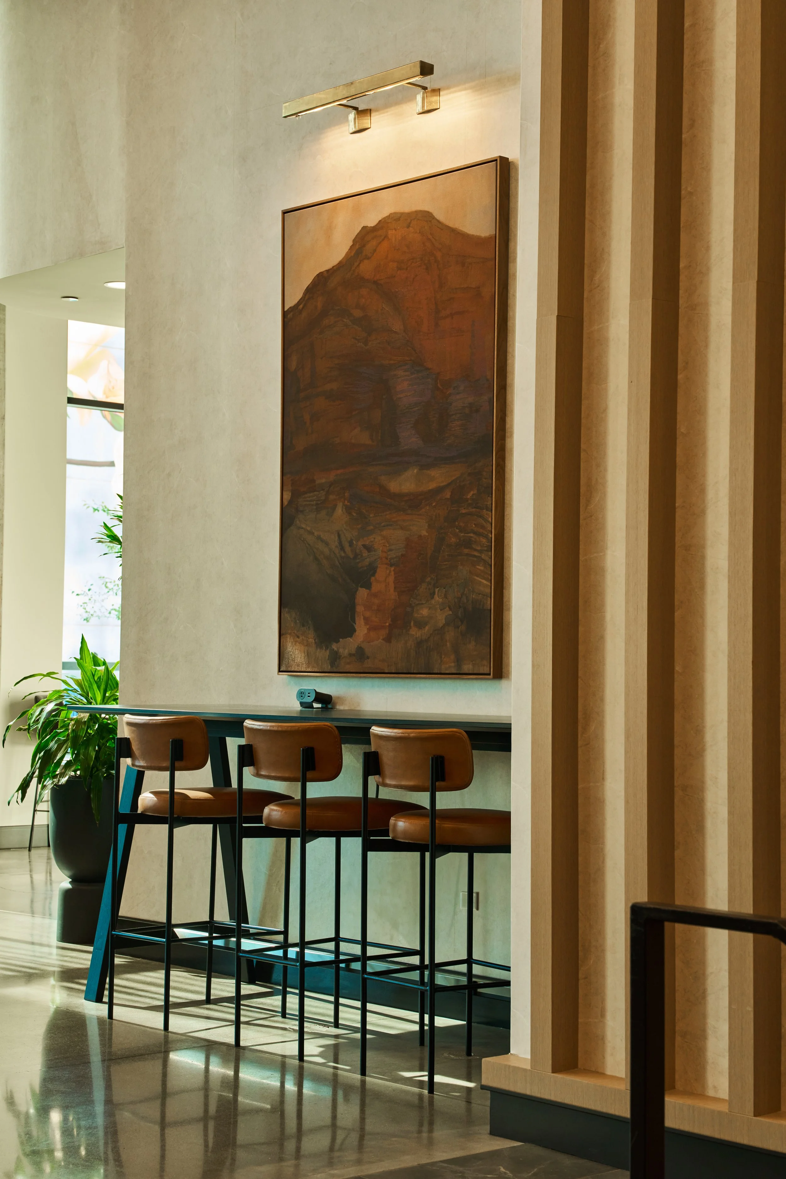 Interior of a modern building with a large painting of a mountain on the wall, a row of brown leather chairs, a potted plant, and sunlight coming through windows.