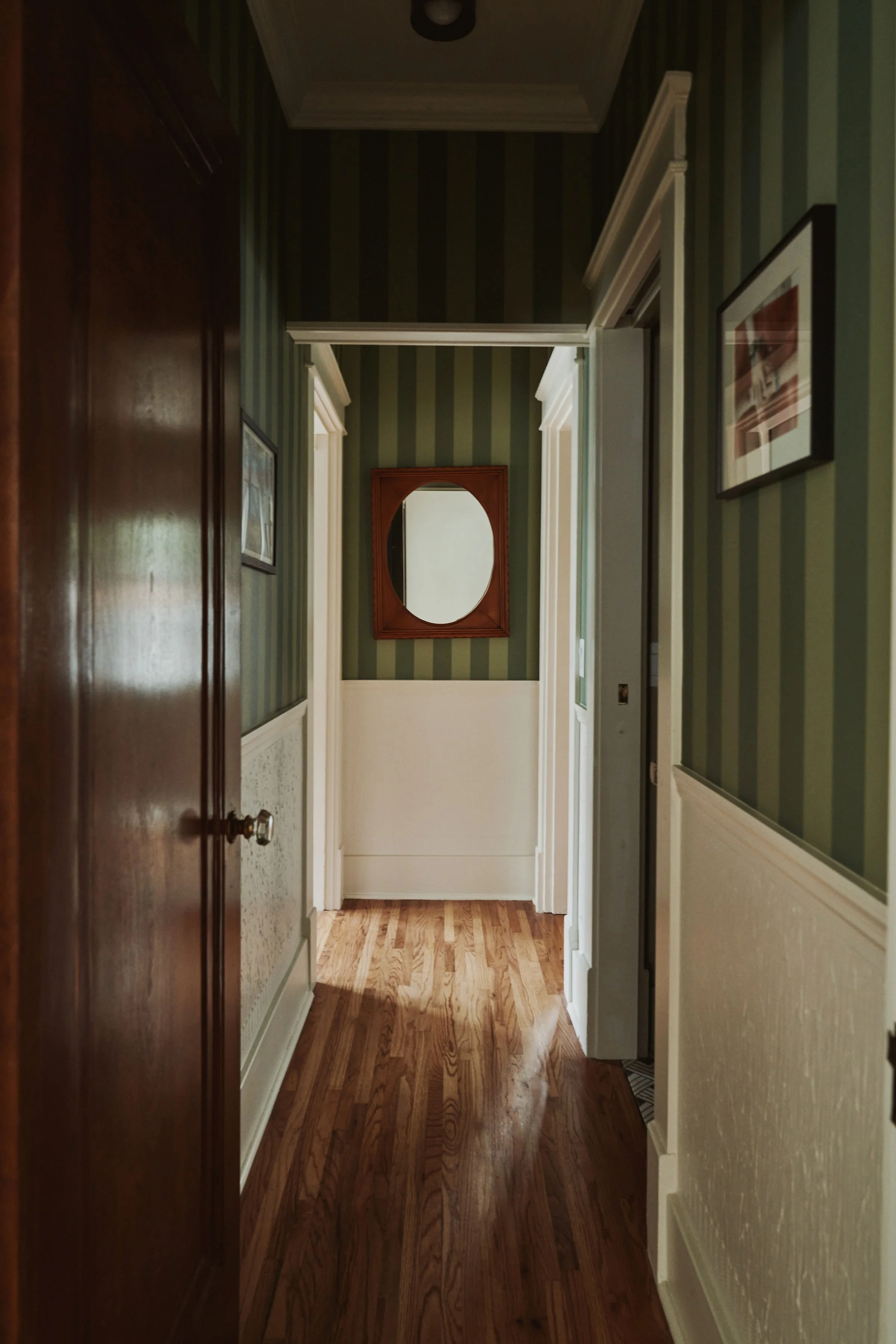 A hallway with hardwood flooring and striped green wallpaper, featuring framed pictures on the walls and an oval mirror at the end.