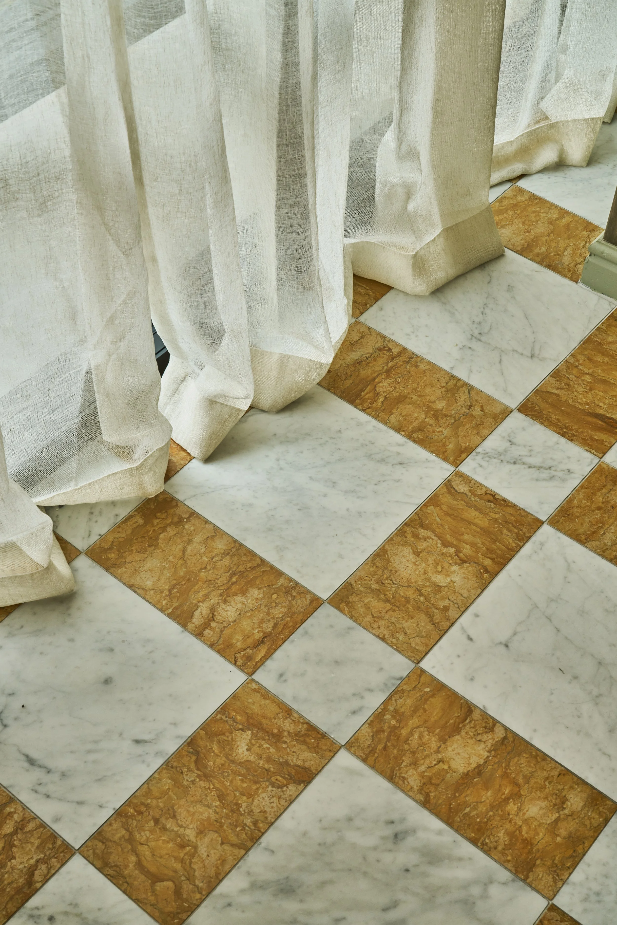 Close-up of a floor with white and brown marble tiles arranged in a pattern. Sheer curtains are hanging and partially covering the tiles.