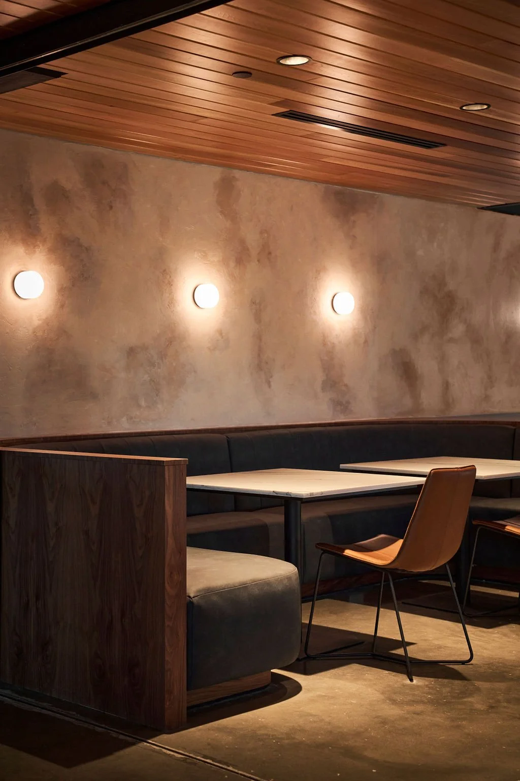 Interior shot of a modern restaurant or café with a textured beige wall, wooden ceiling, wall-mounted lights, a black cushioned bench, and a wooden chair next to a white table.