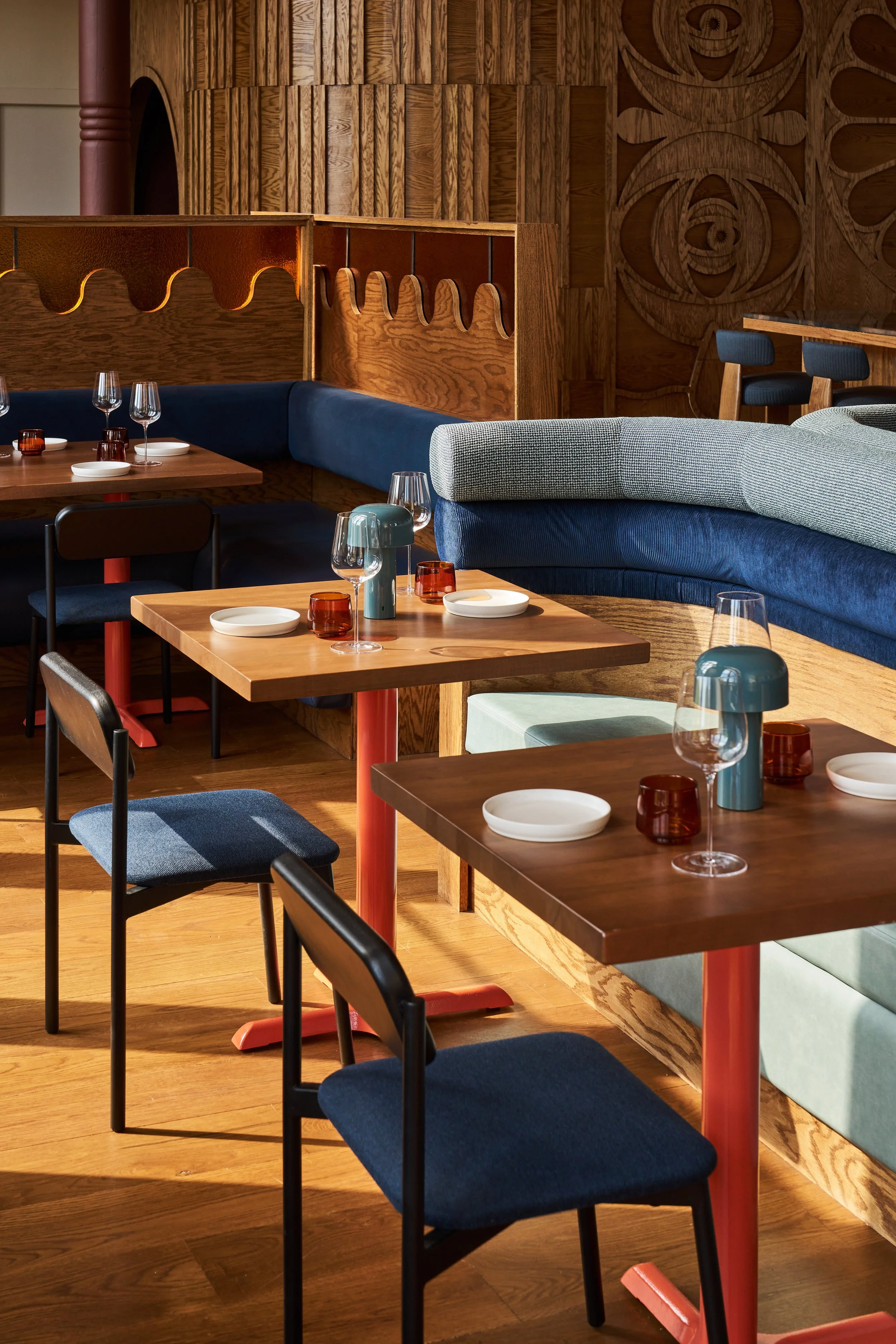 Interior of a restaurant featuring wooden tables, blue and gray cushioned chairs, a blue upholstered banquette, and a wooden partition with decorative cutouts, with a large wooden wall with carved designs in the background.