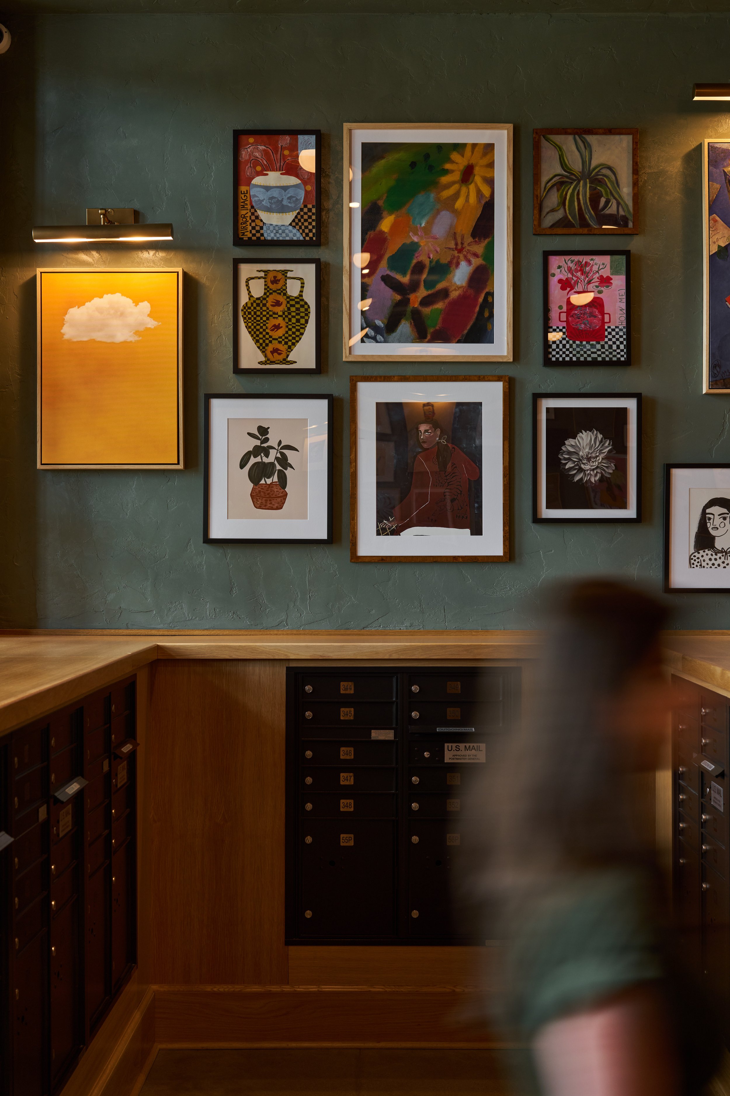 Interior of a building with a green wall decorated with multiple framed artworks, including paintings and illustrations of vases, plants, and a woman. There are mailboxes on the lower part of the image and a wooden counter or shelf in front. The ligh