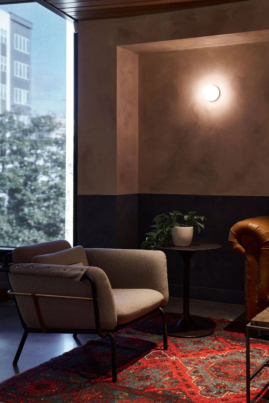 An interior scene with a beige armchair, a small round table with a potted plant, a textured wall with a round wall sconce, and a large window showing trees and a building outside.