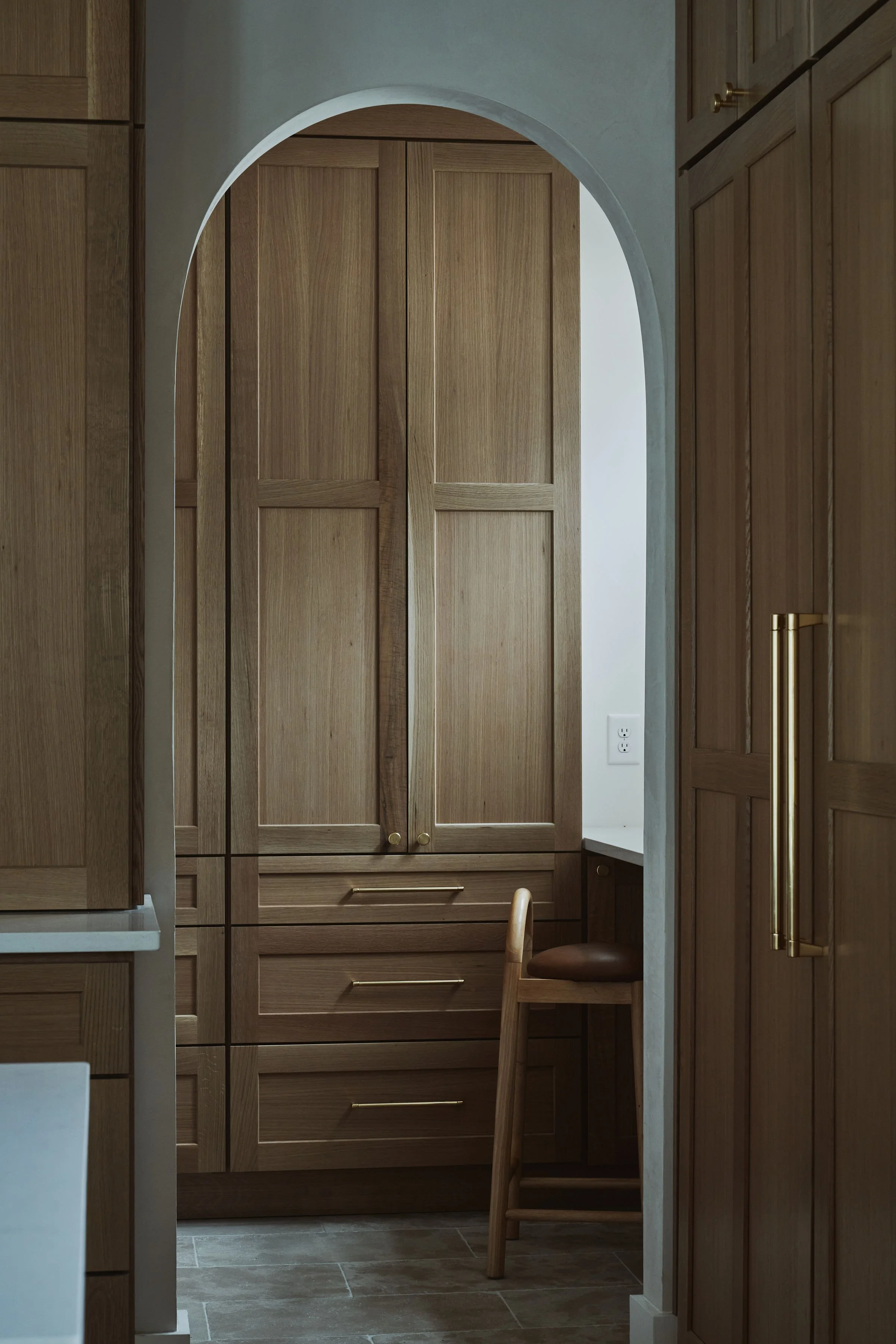 View of a wooden kitchen with cabinets, a chair, and a small desk, seen through an arched entryway.