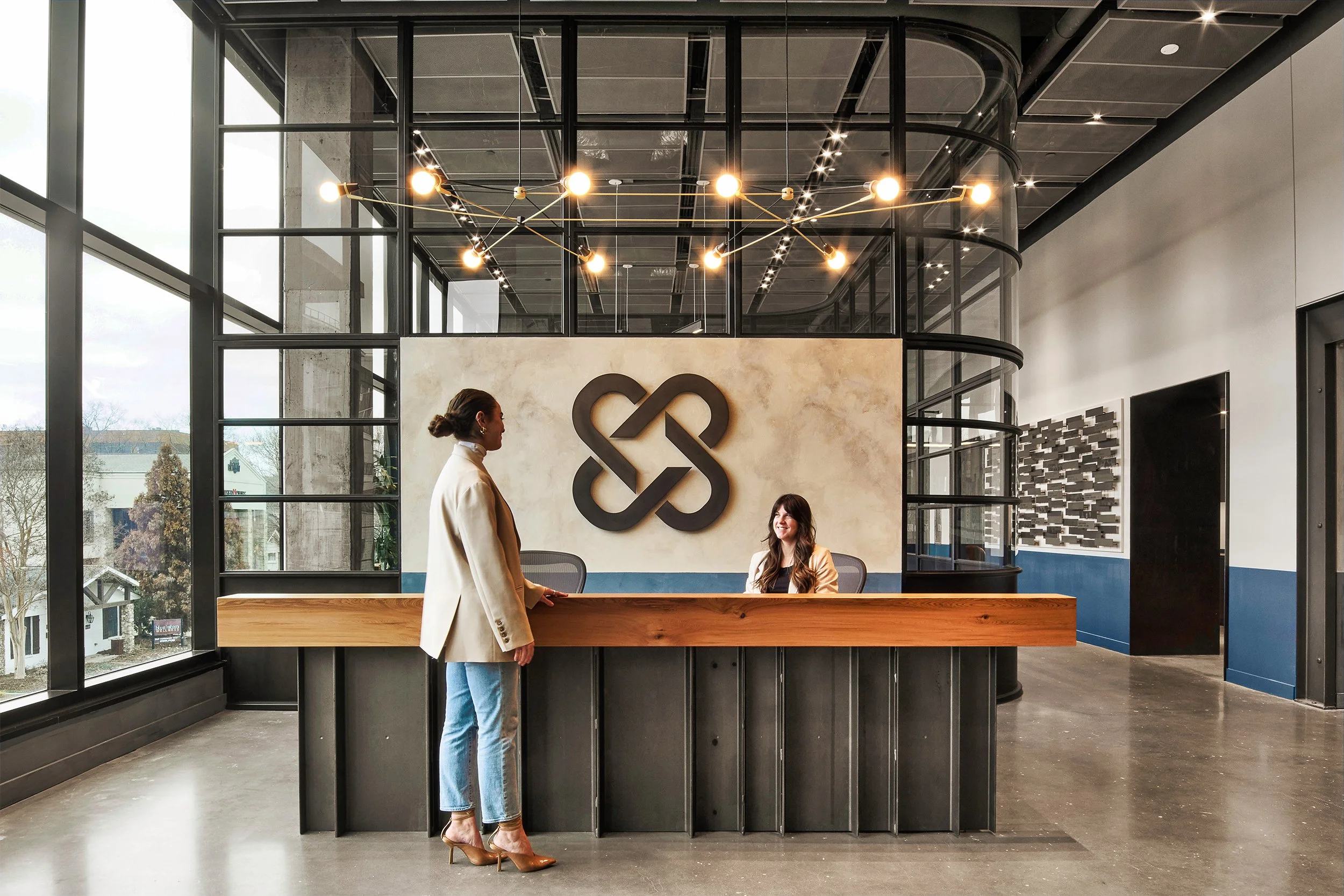 Modern interior of an office lobby with a reception desk, two women, one standing and one sitting, engaged in conversation, large windows, an abstract wall logo, and a chandelier overhead.